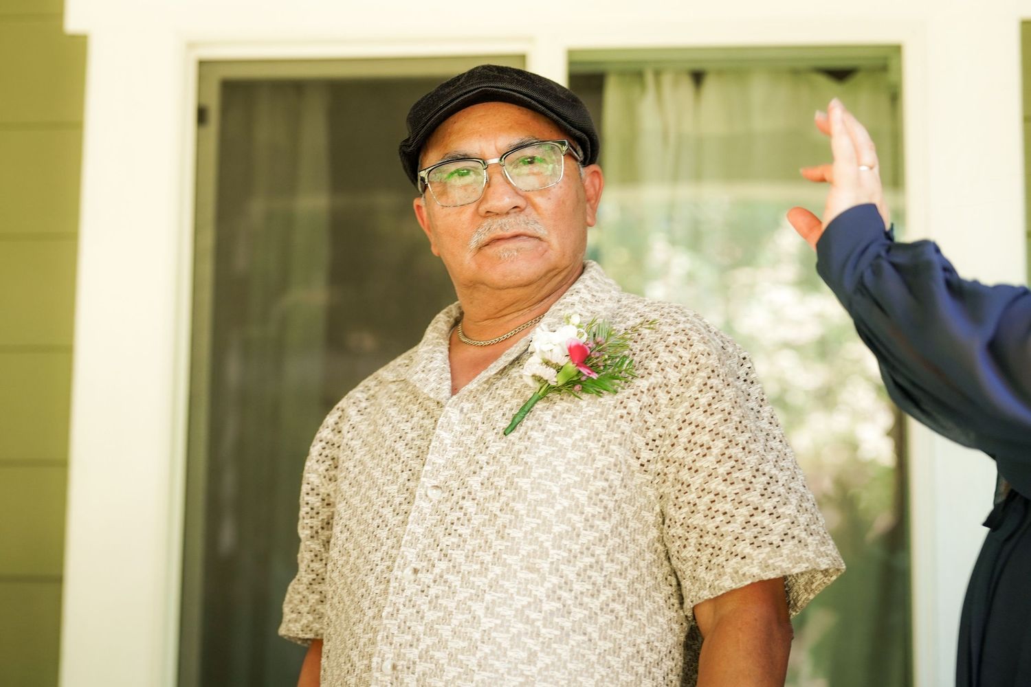 Older man in a flat cap and patterned shirt wears a pink and white boutonniere while standing outside before a wedding.