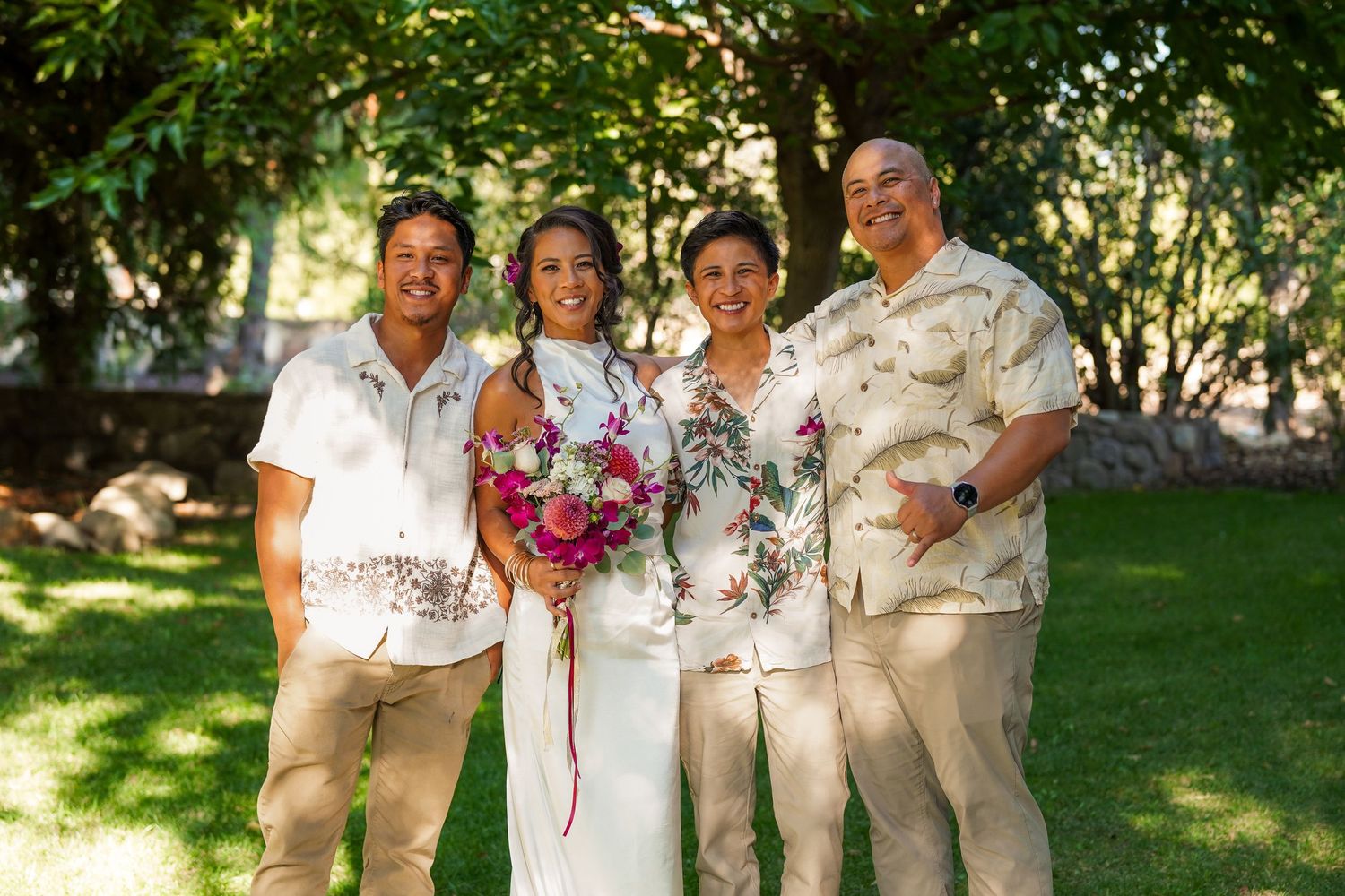 A bride holding a vibrant pink bouquet poses with three men in matching floral Hawaiian shirts in a sunny garden wedding portrait.