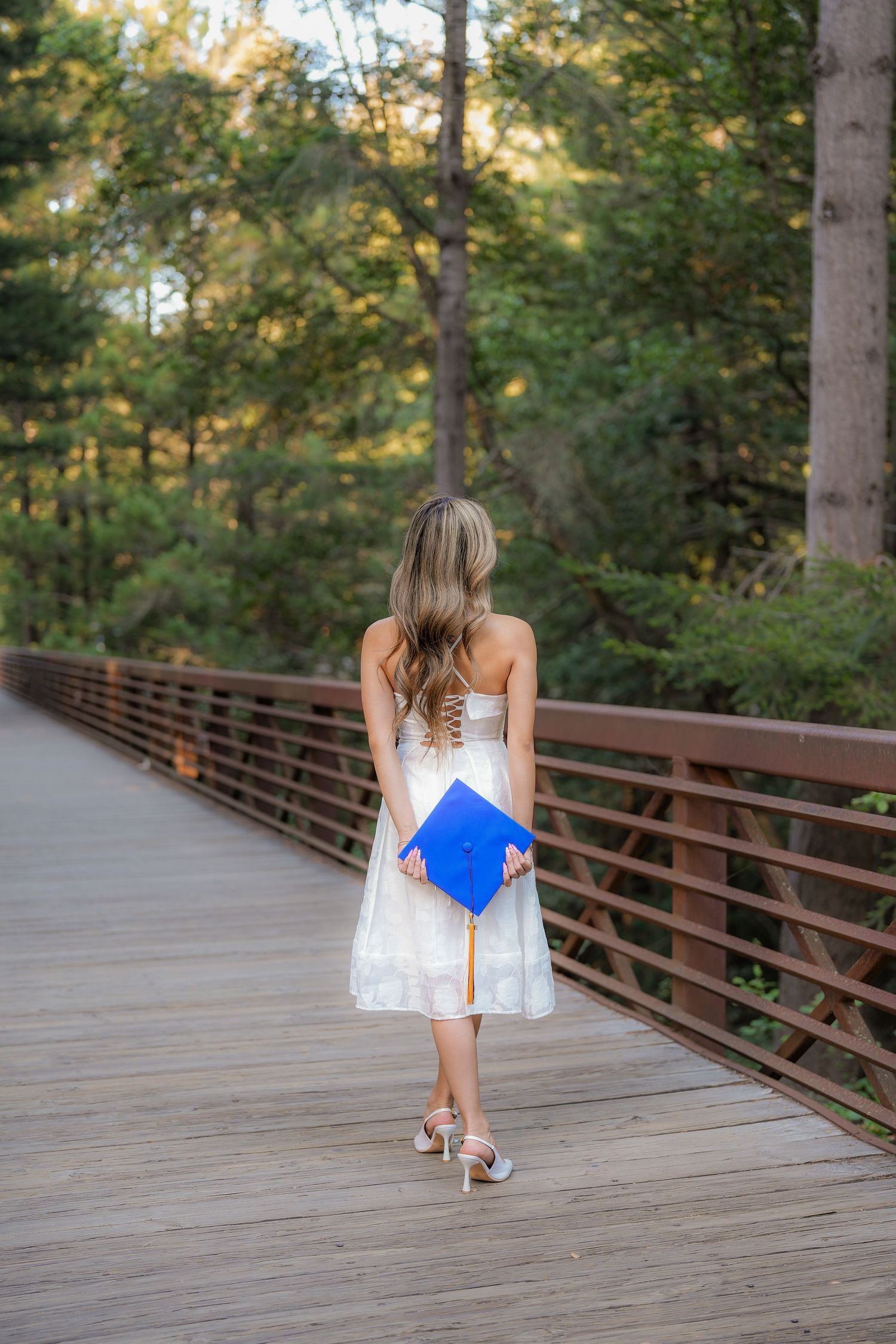 Graduate in a white dress walks away on a wooden forest bridge, holding a blue cap with a gold tassel behind her back.