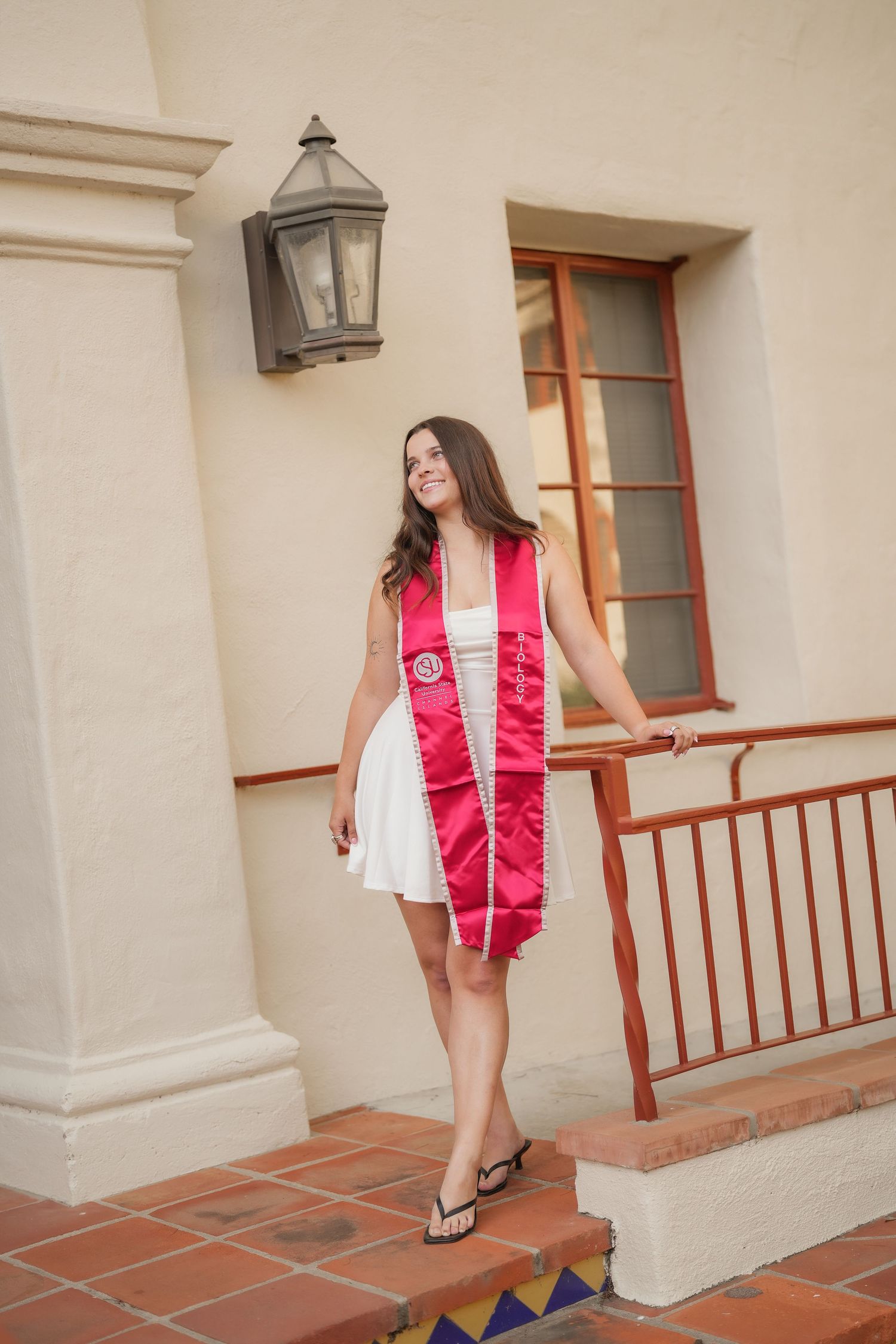 Smiling graduate in a white dress and pink graduation stole poses beside a stucco building with a lantern and red tile steps.