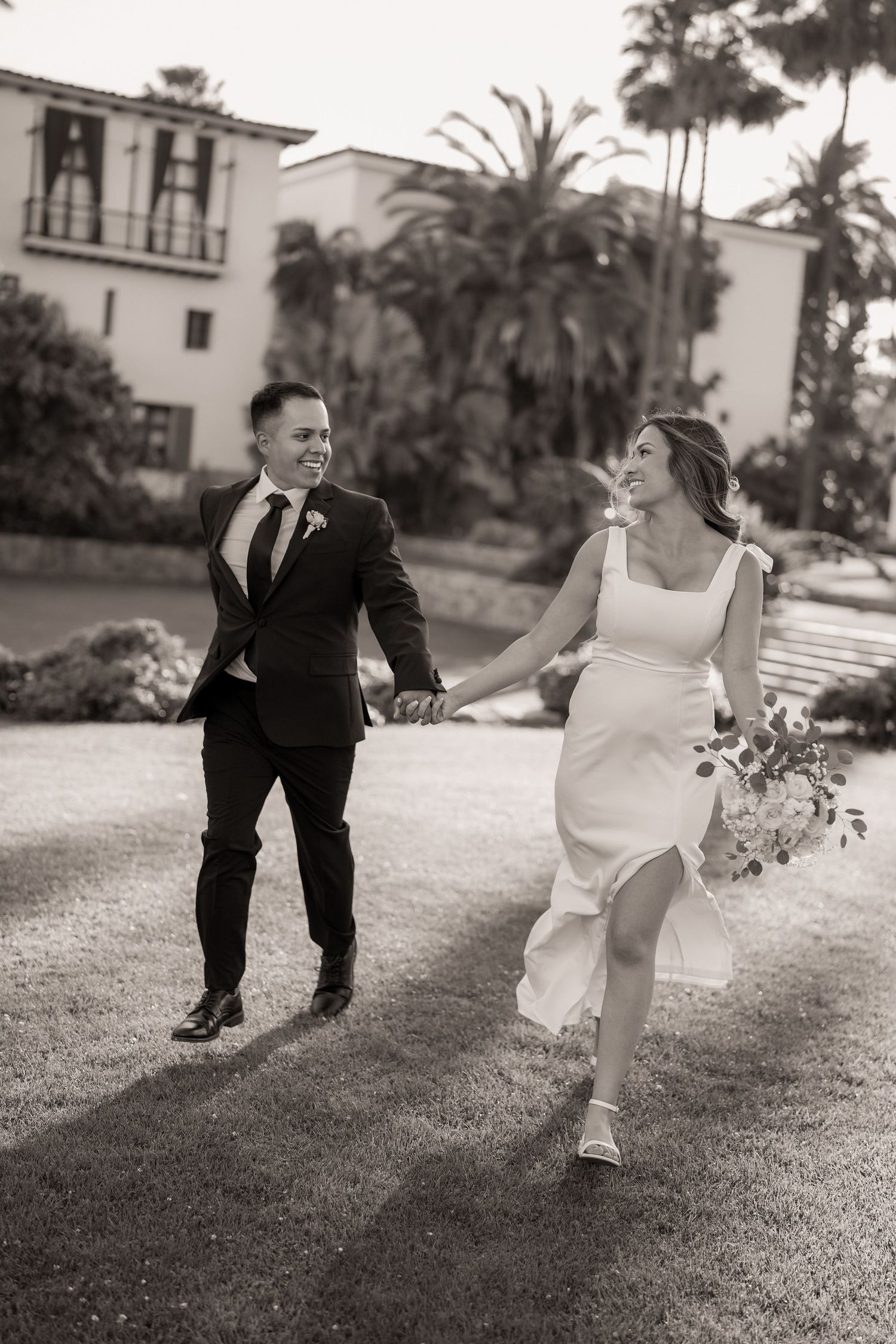 Smiling bride holding a bouquet and groom in a dark suit run hand in hand across a sunlit lawn in a black and white photo.