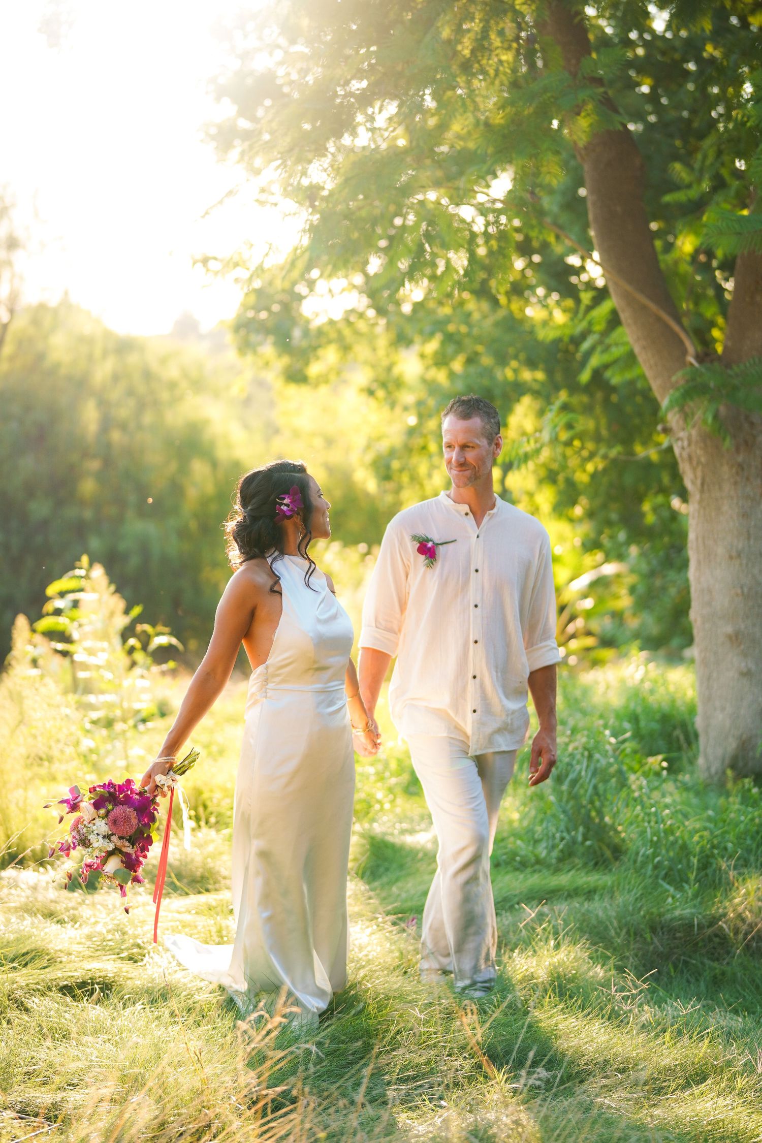 Smiling couple in wedding attire holding hands and walking through a sunlit grassy field with a pink bouquet.