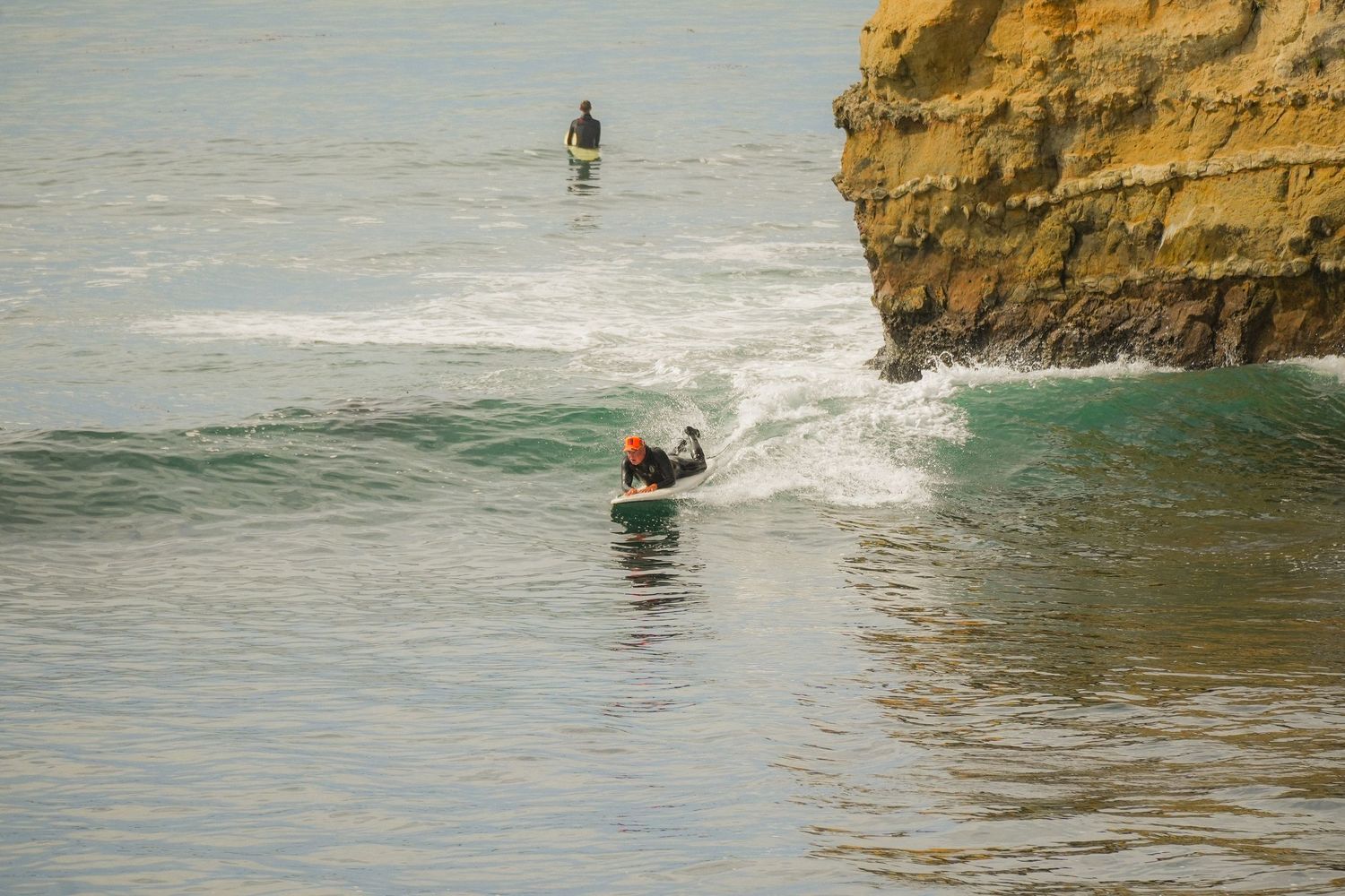 Surfer lies on a board in the water near a golden sandstone cliff while another surfer stands in the distance.