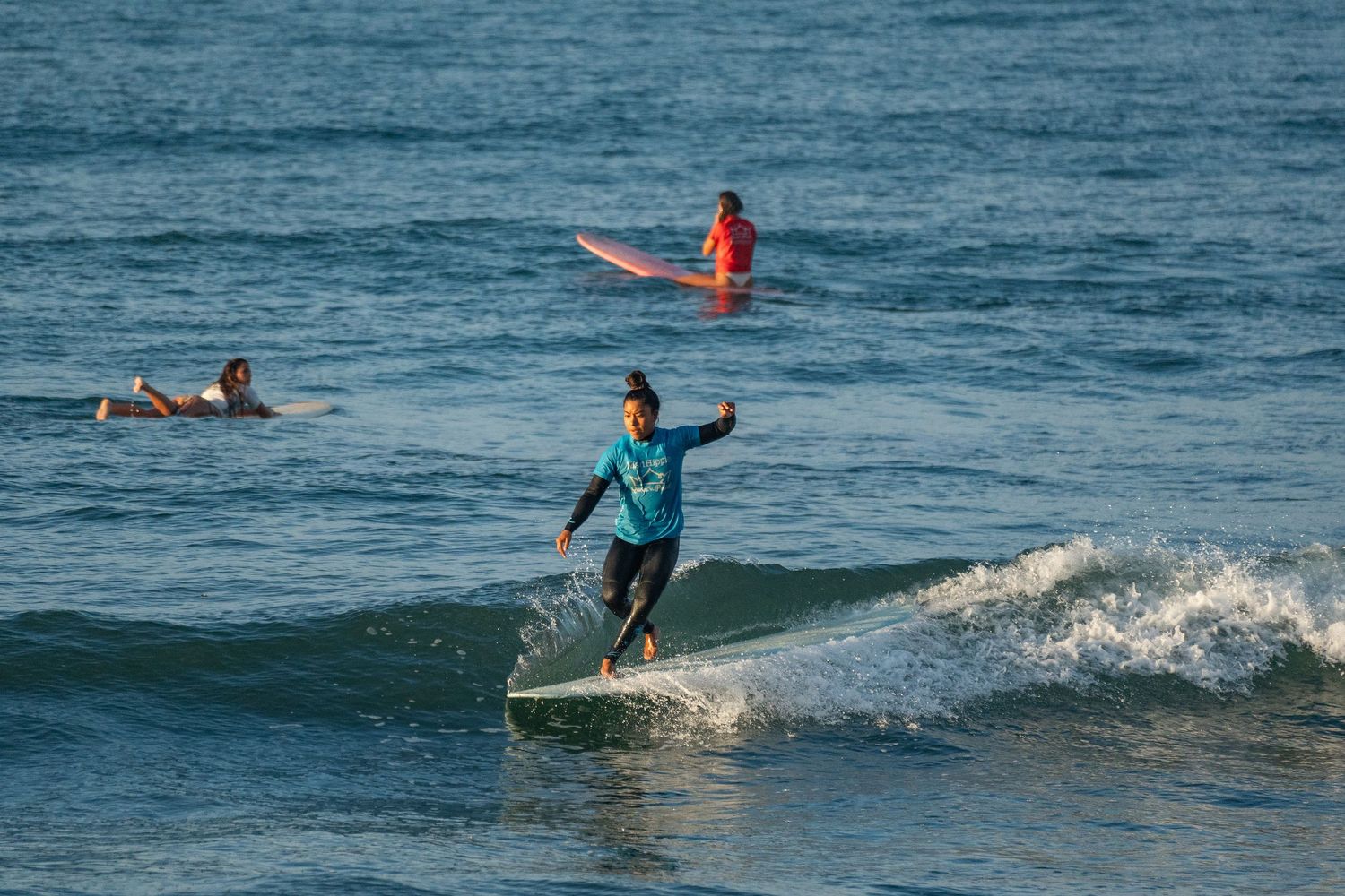 Surfer in a teal shirt rides a wave on a longboard while two other surfers wait in the ocean behind.