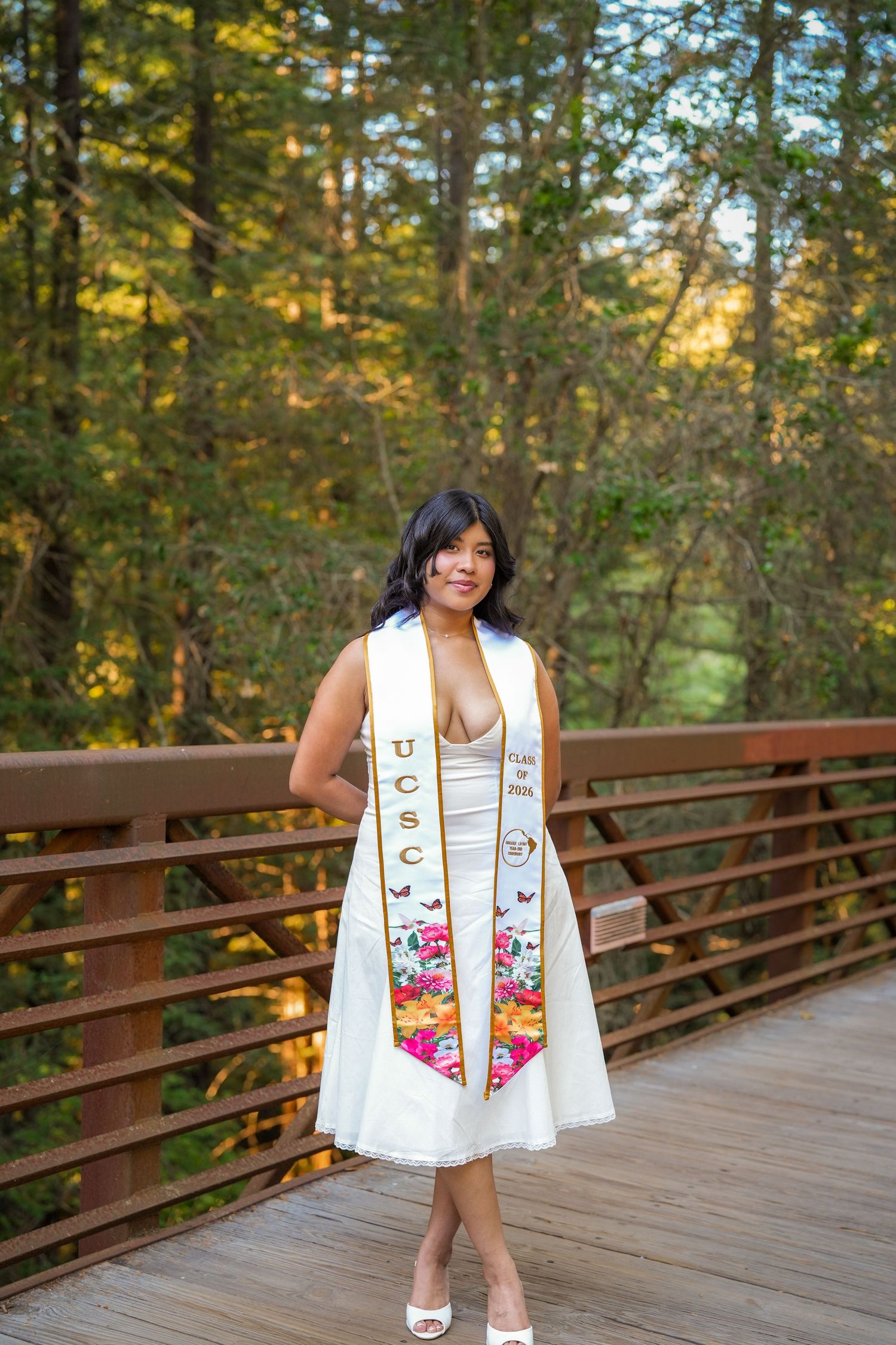 Smiling graduate in a white dress and floral UCSC graduation stole poses on a wooden bridge surrounded by tall redwood trees.