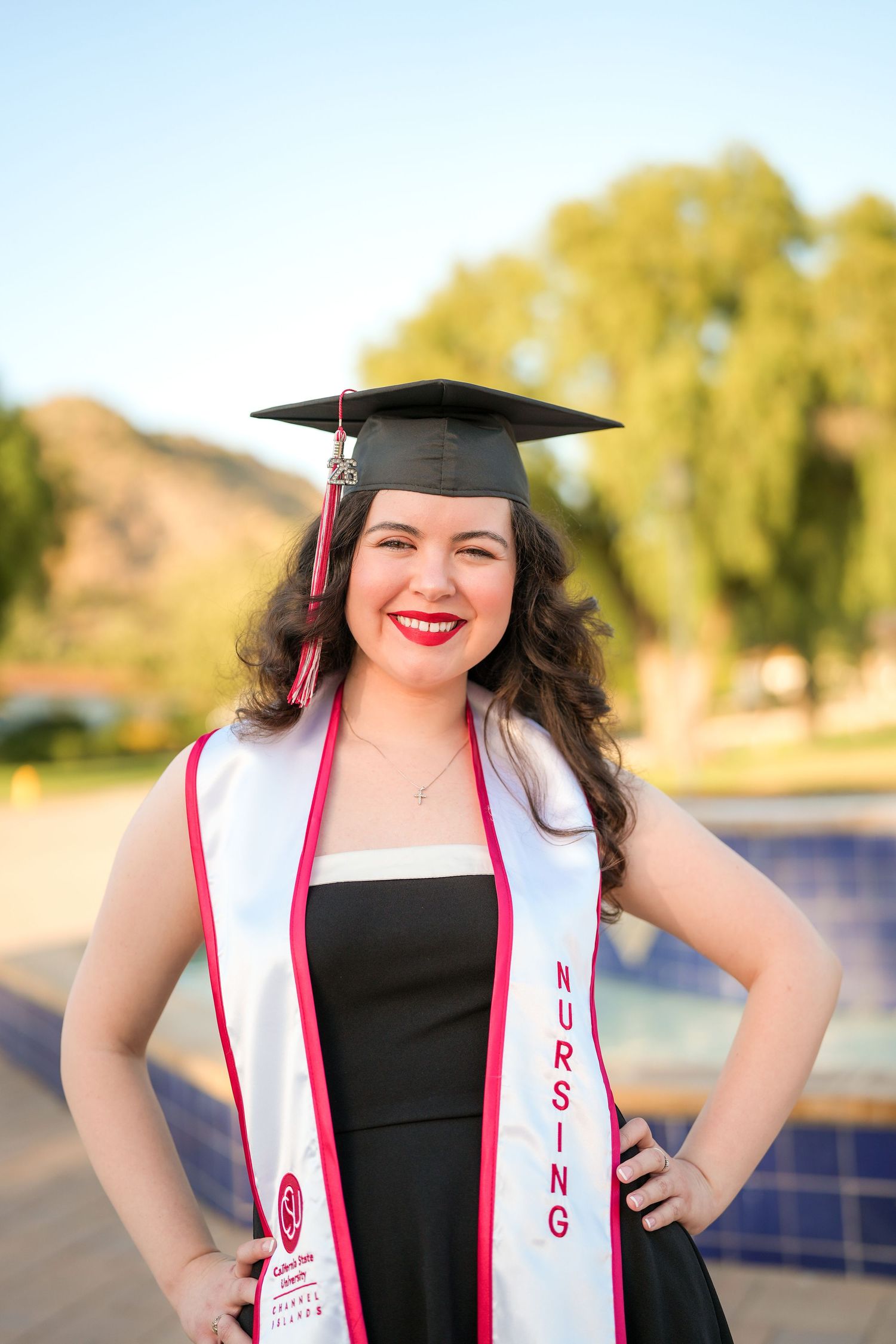 A close-up of a smiling nursing graduate with red lipstick, a black cap, and a white nursing stole standing beside a tiled fountain.