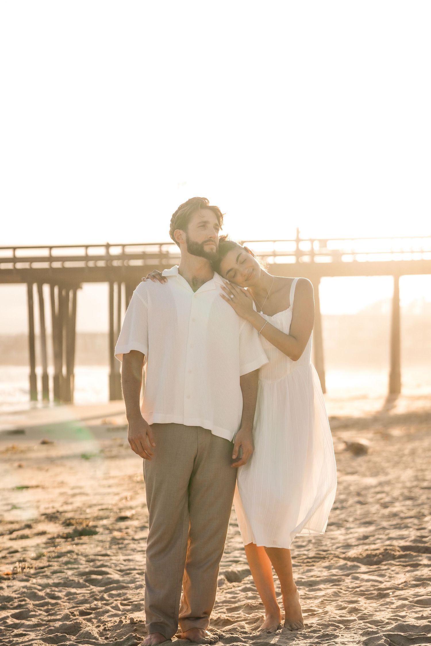 Couple in white standing close on a sunlit beach at golden hour with a wooden pier in the background.