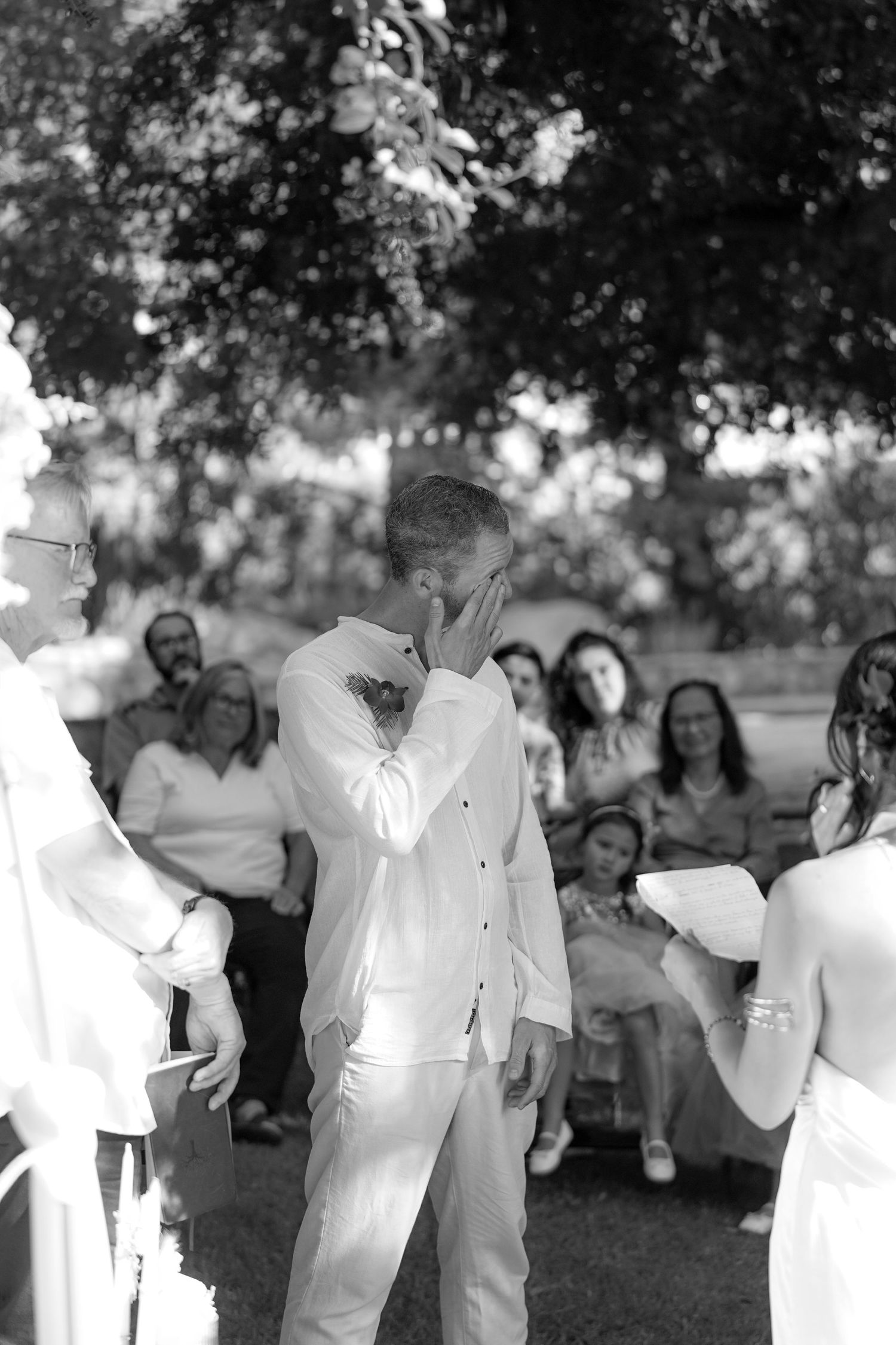 In a black and white photo, a groom wipes his face with emotion as the bride and guests watch during an outdoor wedding ceremony.