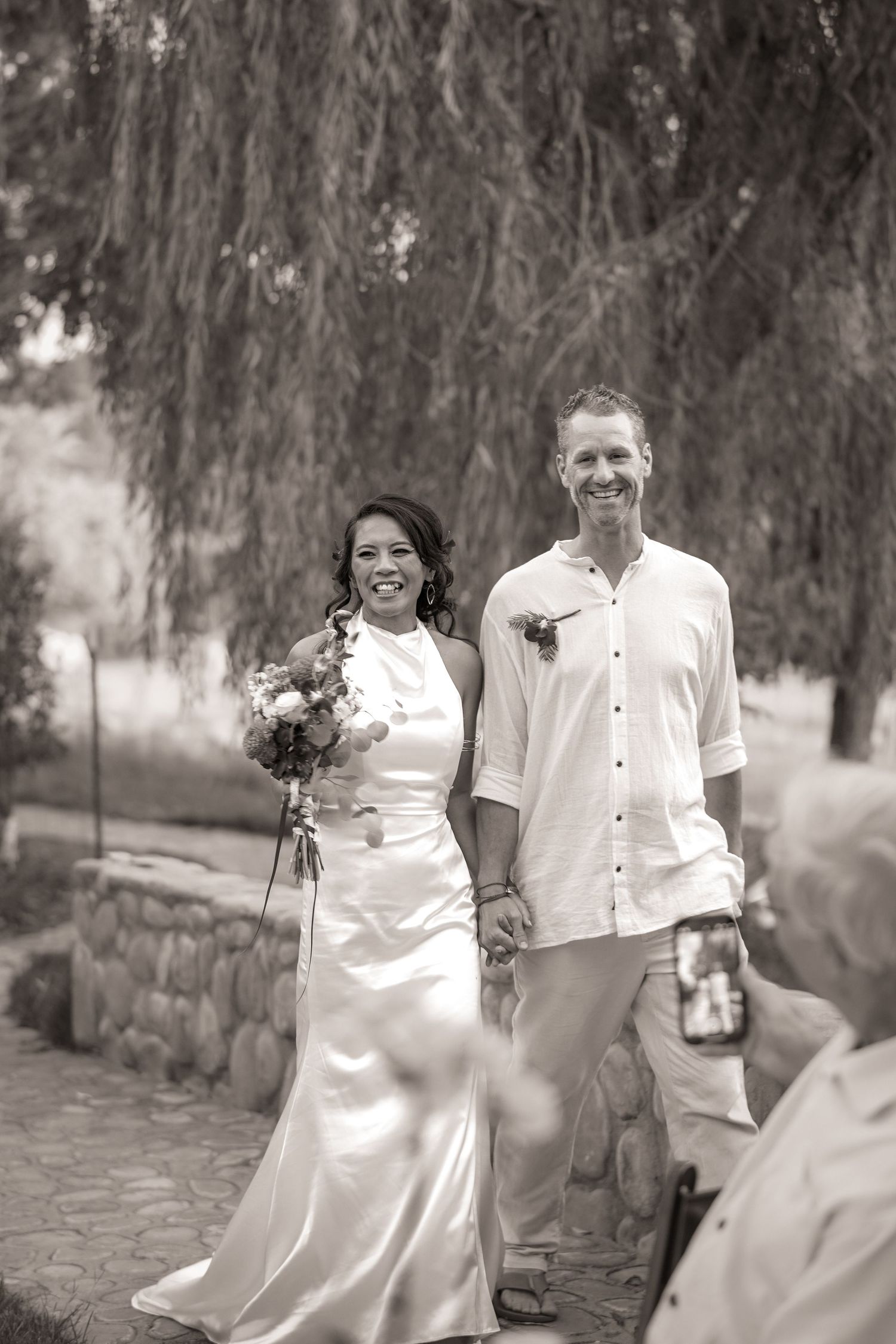 Black and white photo of a joyful bride and groom walking hand in hand down the aisle as a guest films with a phone.