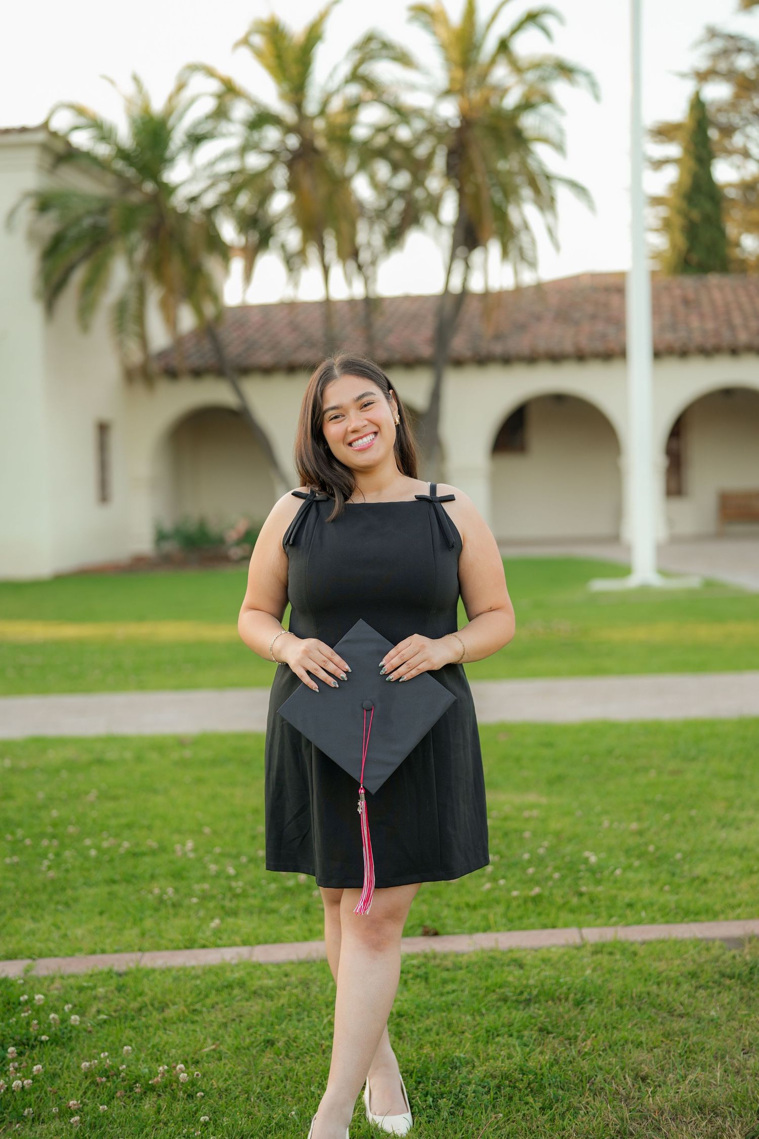 A graduate in a black dress smiles while holding her cap with a pink tassel on a green lawn beside palm trees and a campus building.