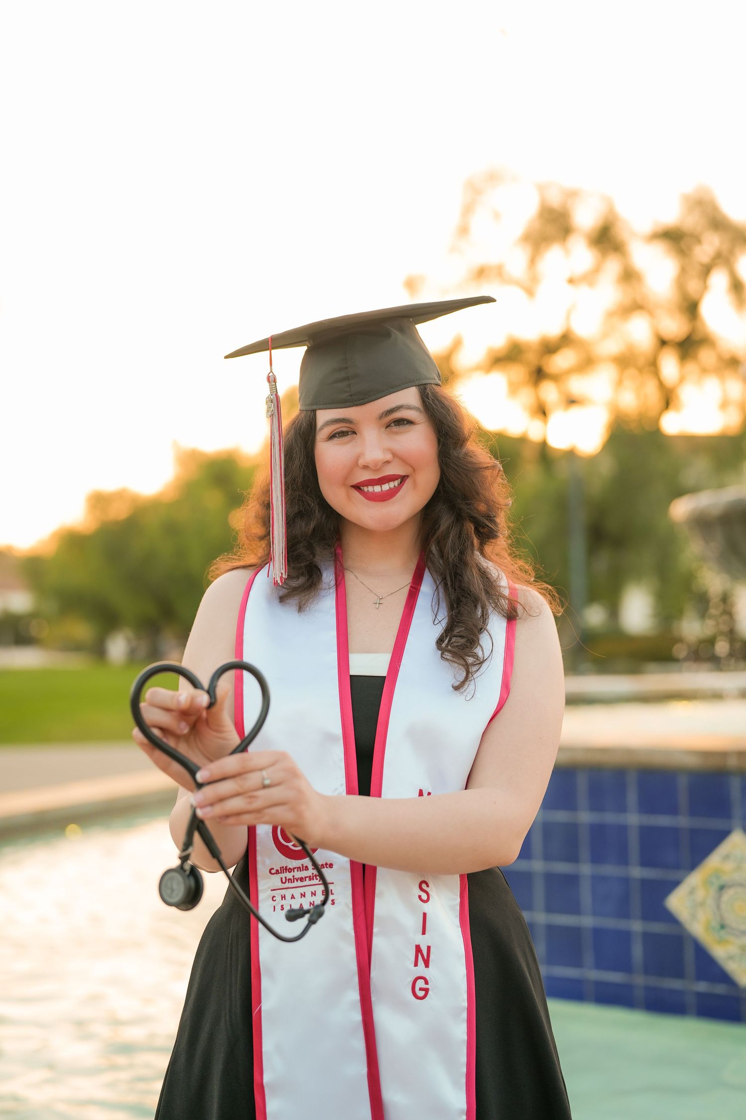 Smiling nursing graduate in cap and sash shapes her stethoscope into a heart beside a sparkling blue-tiled fountain.