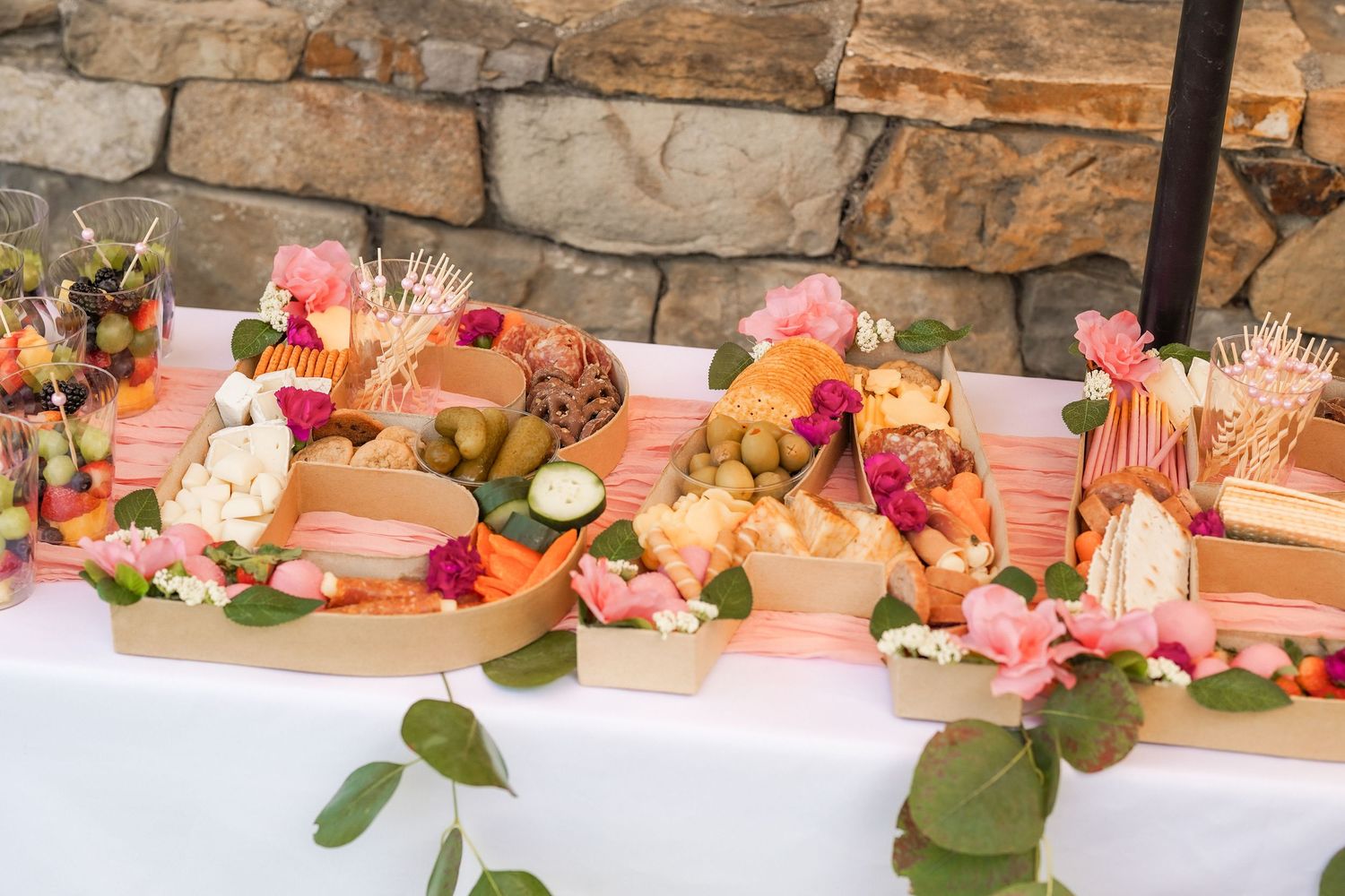 Elegant grazing table with cheeses, fruit, crackers, and fresh pink flowers on a white tablecloth against a rustic stone wall.