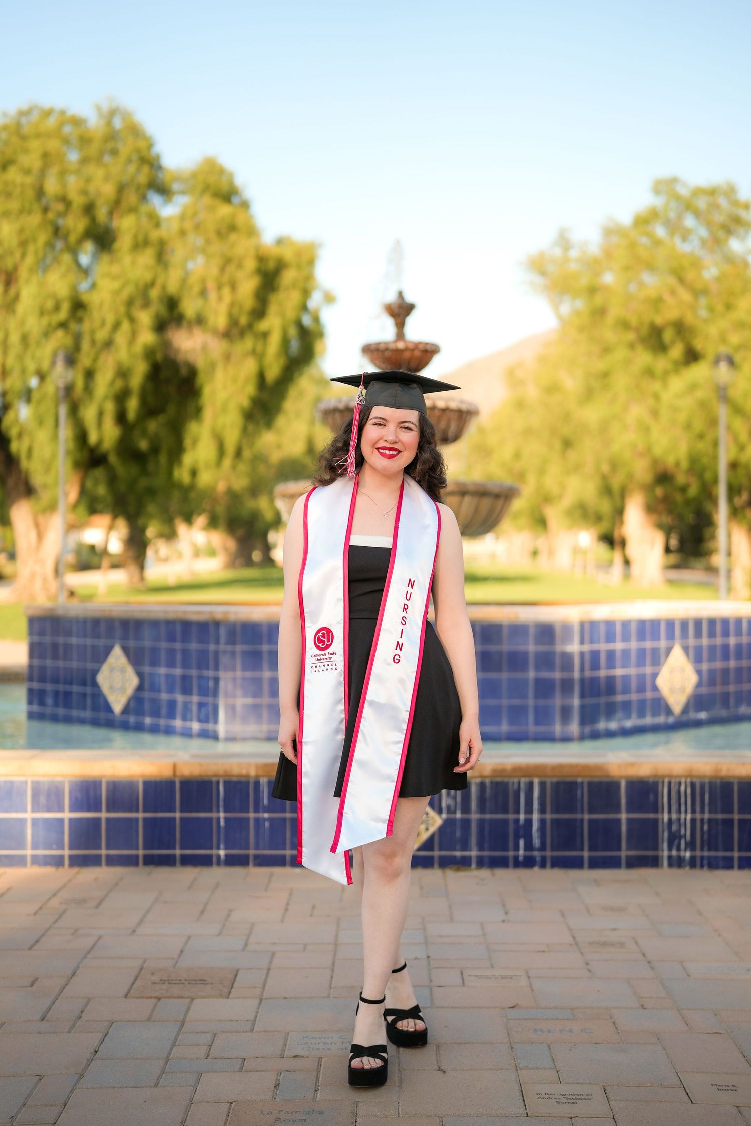 Smiling nursing graduate in a cap, black dress and white sash poses in front of a blue tiled fountain on graduation day.