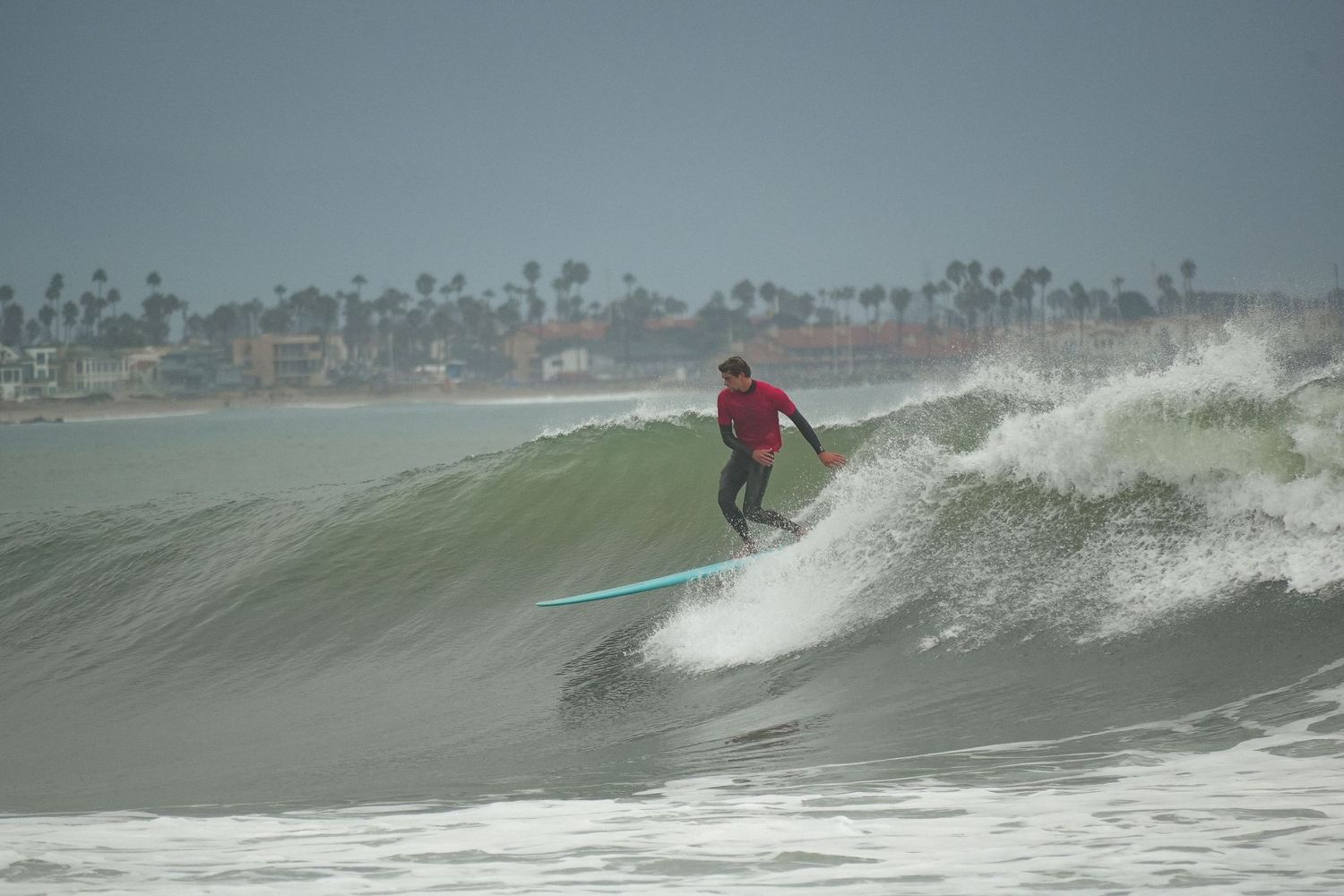 Surfer in a red shirt rides a teal longboard along a breaking wave with palm trees on the shoreline.