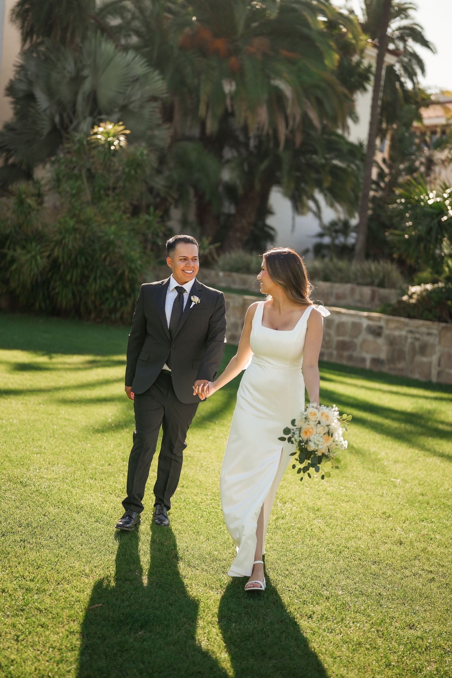 A smiling bride holding a bouquet and a groom in a dark suit hold hands while walking on a sunlit lawn surrounded by palm trees.