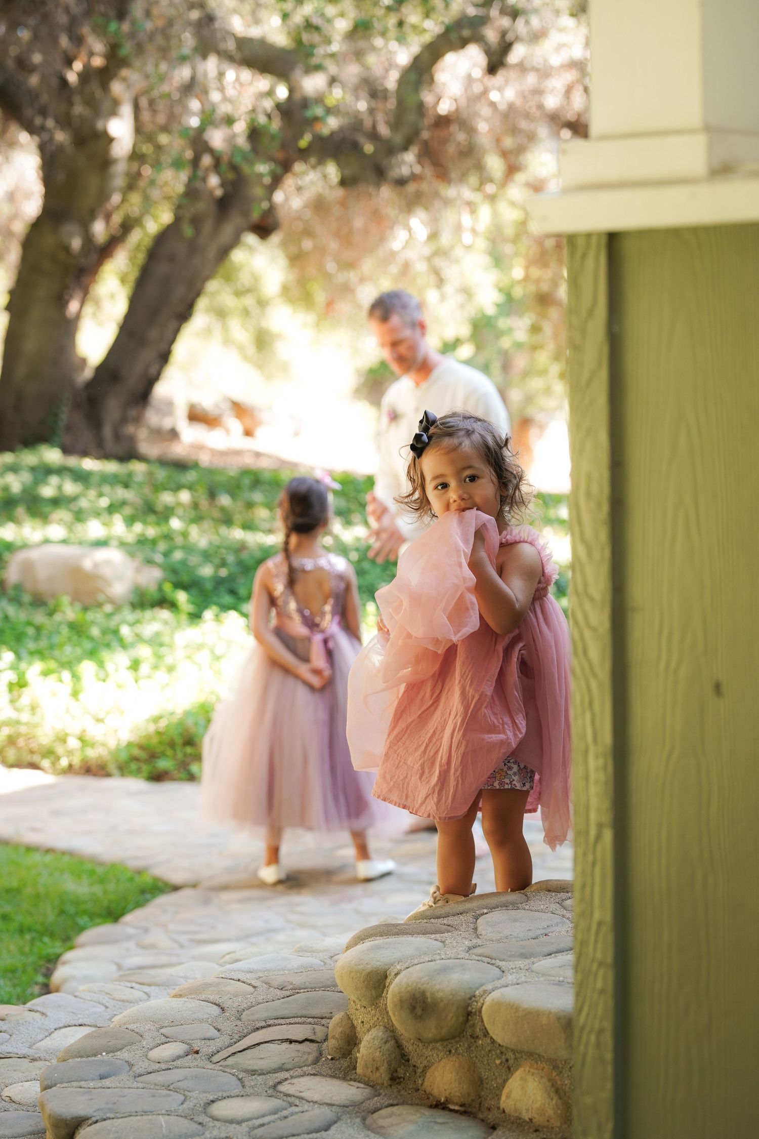 A toddler in a pink tulle dress peeks around a green post at an outdoor wedding while another girl and a man walk along a stone path.