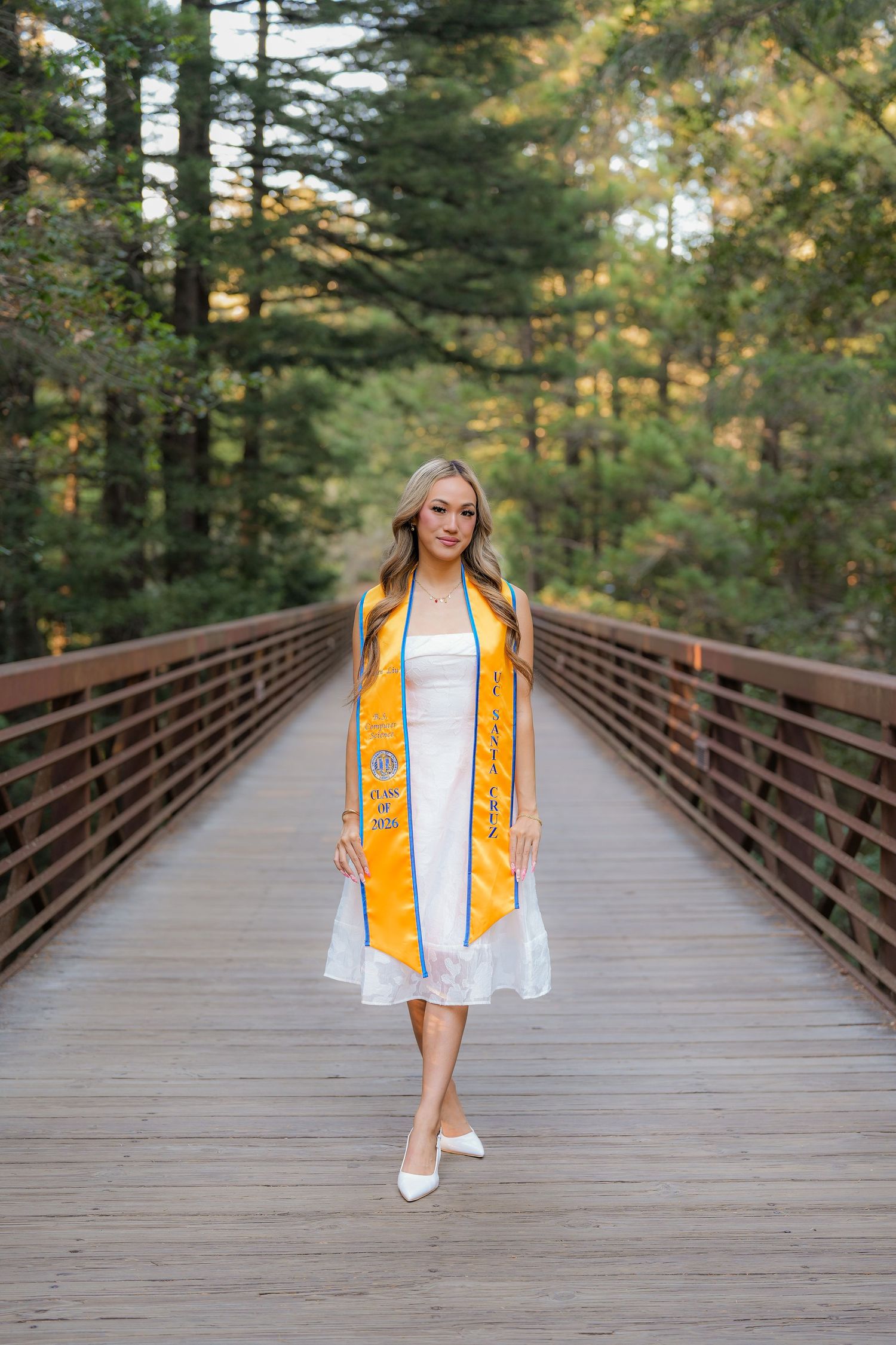 Graduate in a white dress and gold stole walks along a wooden bridge surrounded by tall green trees in a forest.