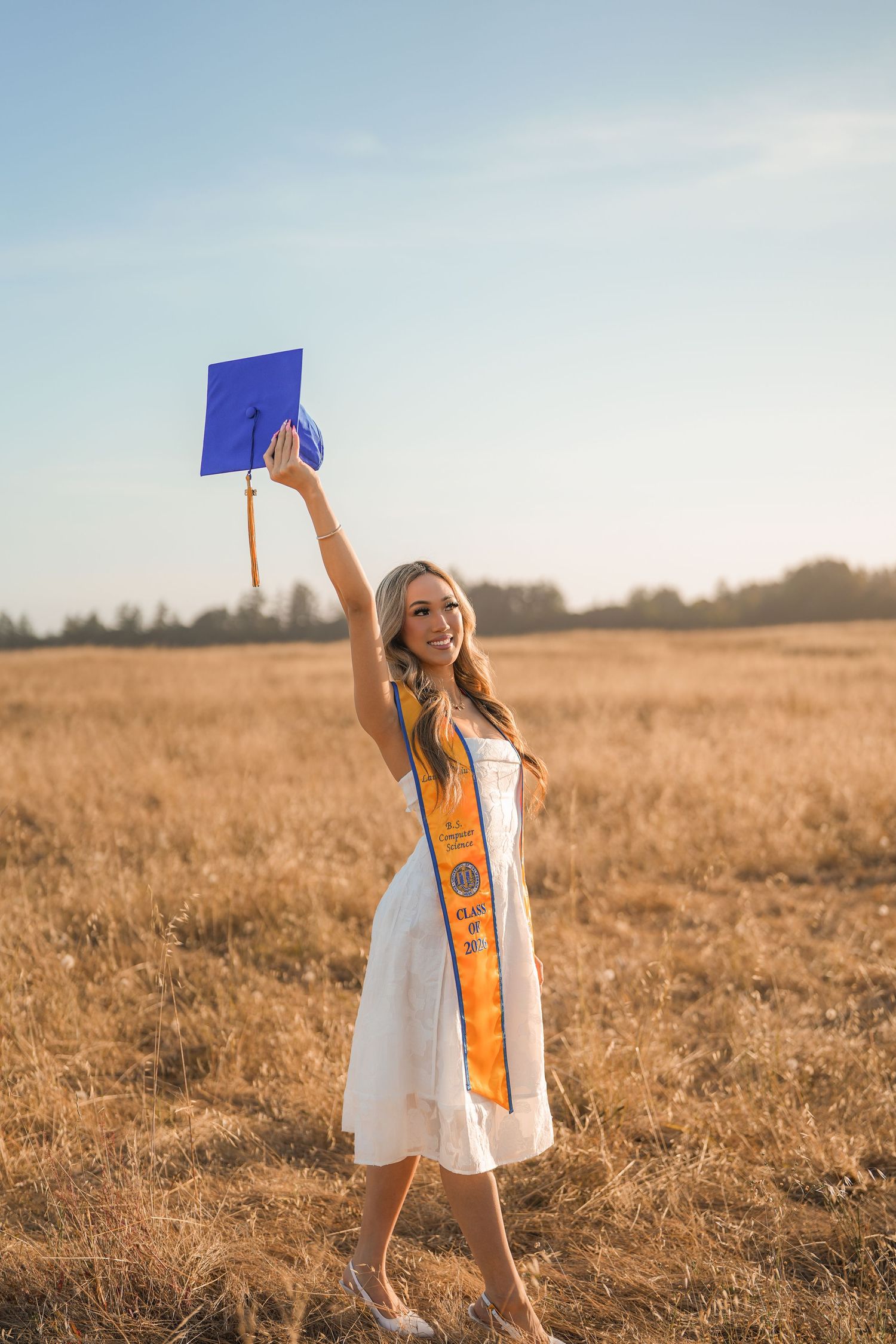 Smiling graduate in an orange stole holds her blue graduation cap high in the air in a sunny golden field.