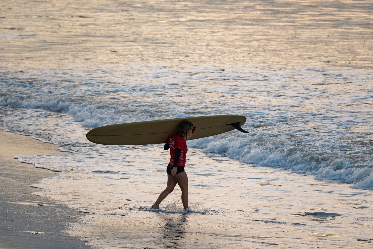 Surfer in a red wetsuit top carries a yellow longboard over her head through shallow water at sunset.