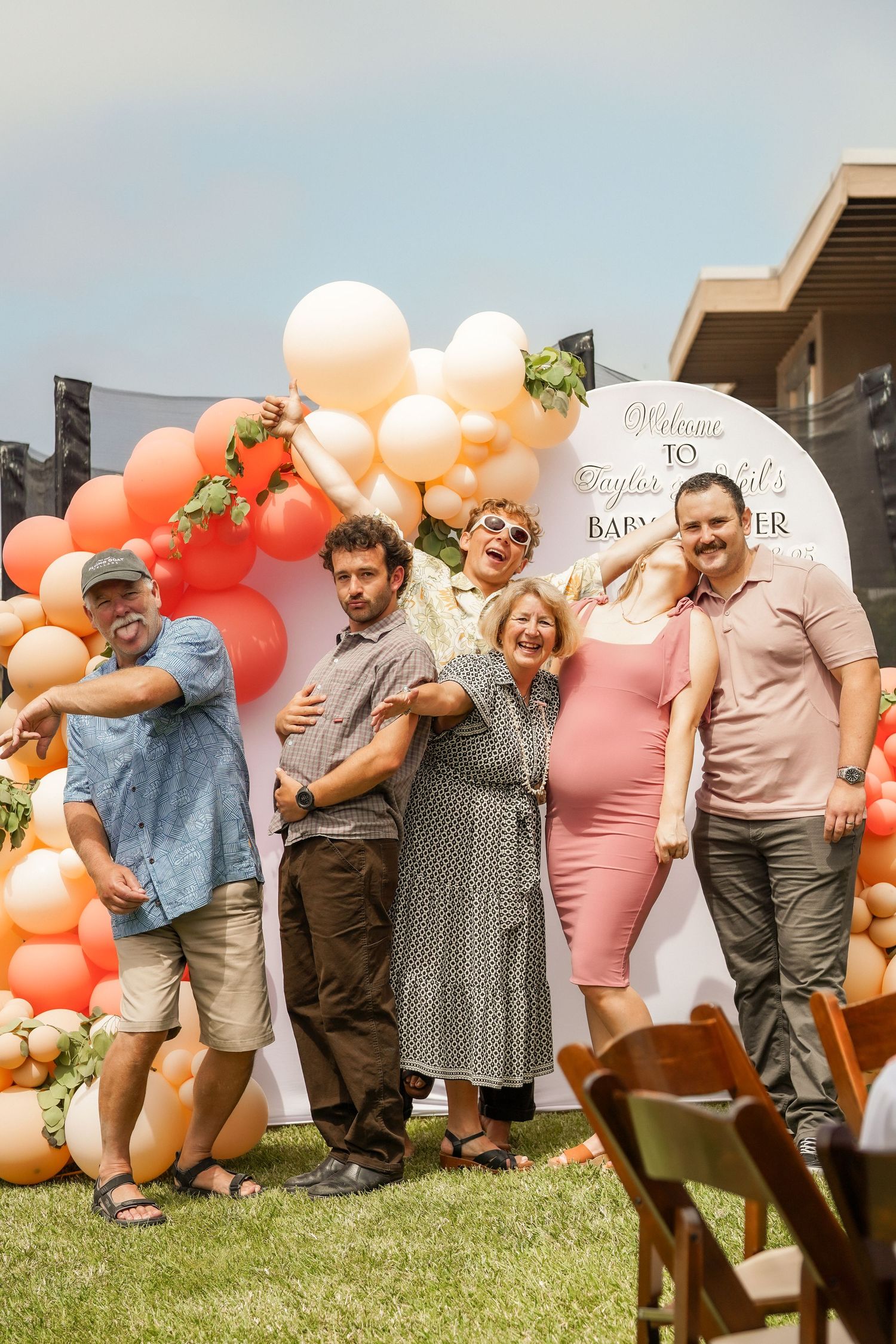 A smiling group of friends and family poses outdoors at a baby shower beside a peach and orange balloon arch and a welcome sign.