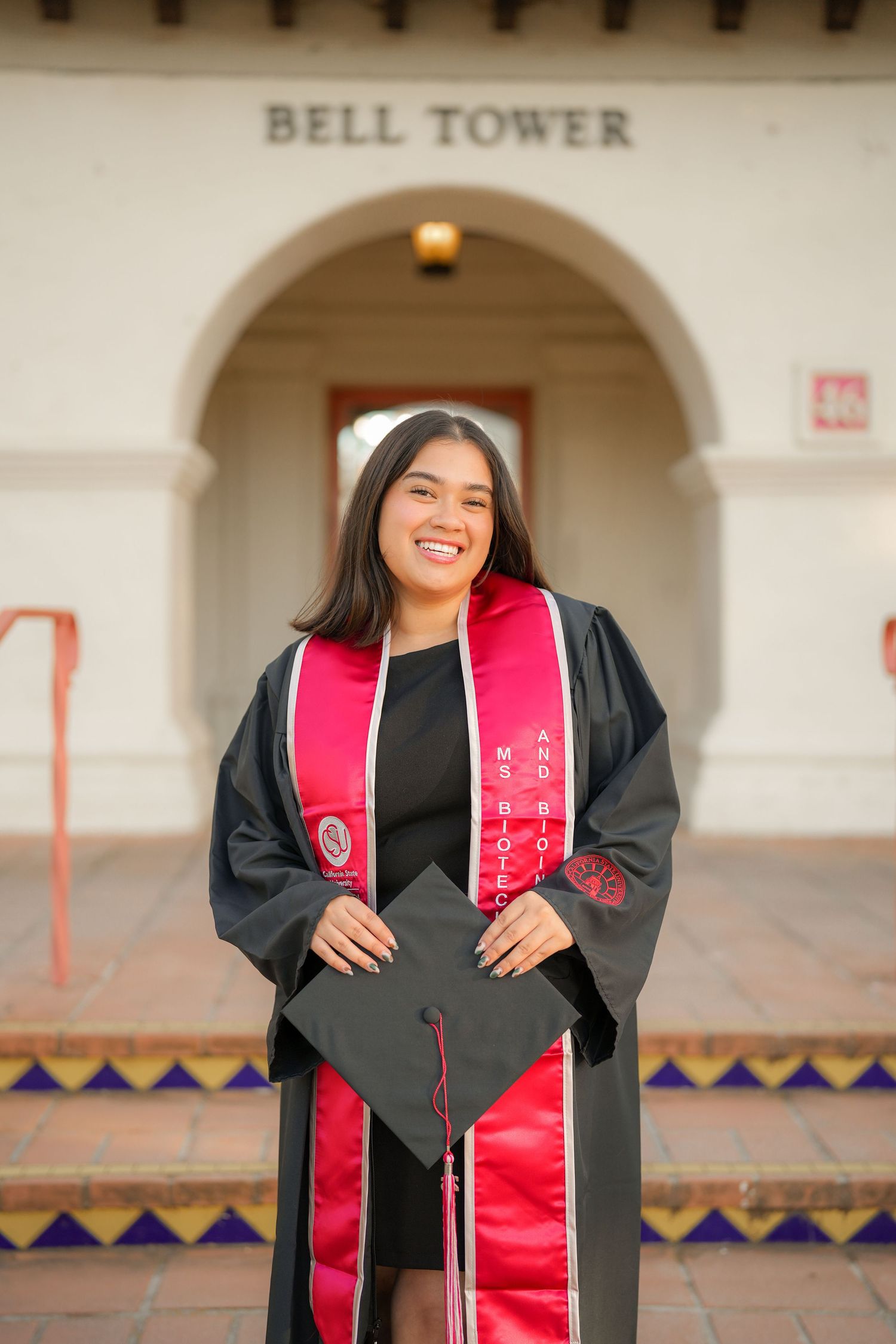 Smiling graduate in black gown and red stole holding her cap in front of the Bell Tower archway on graduation day.