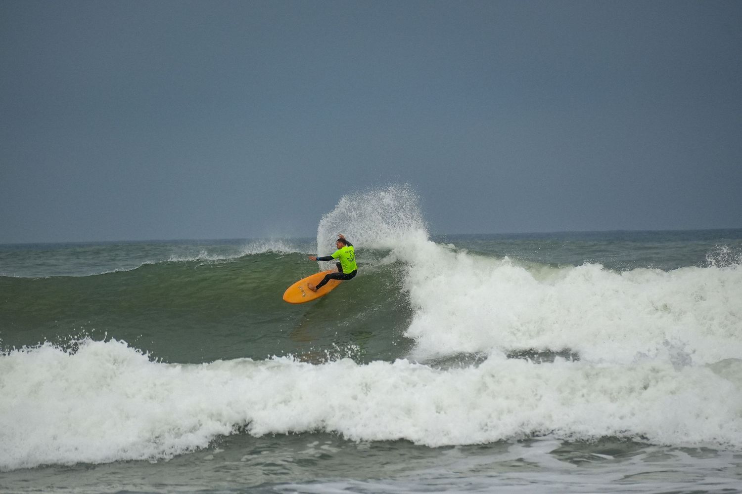 Surfer in a neon green shirt on an orange board makes a sharp turn off the top of a wave, sending spray into the air.
