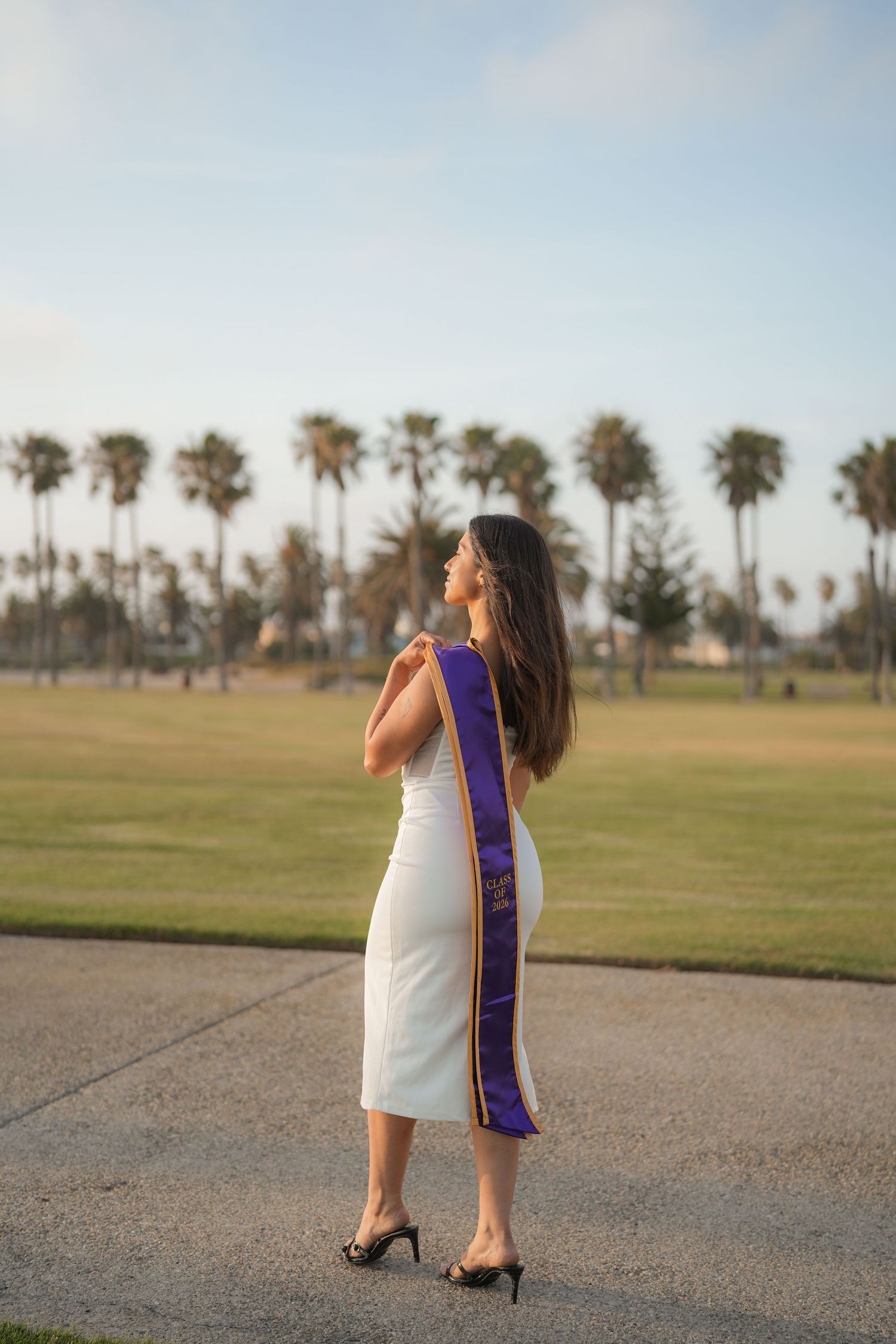 Graduate in a white dress and purple stole poses with her back turned on a coastal path lined with palm trees.