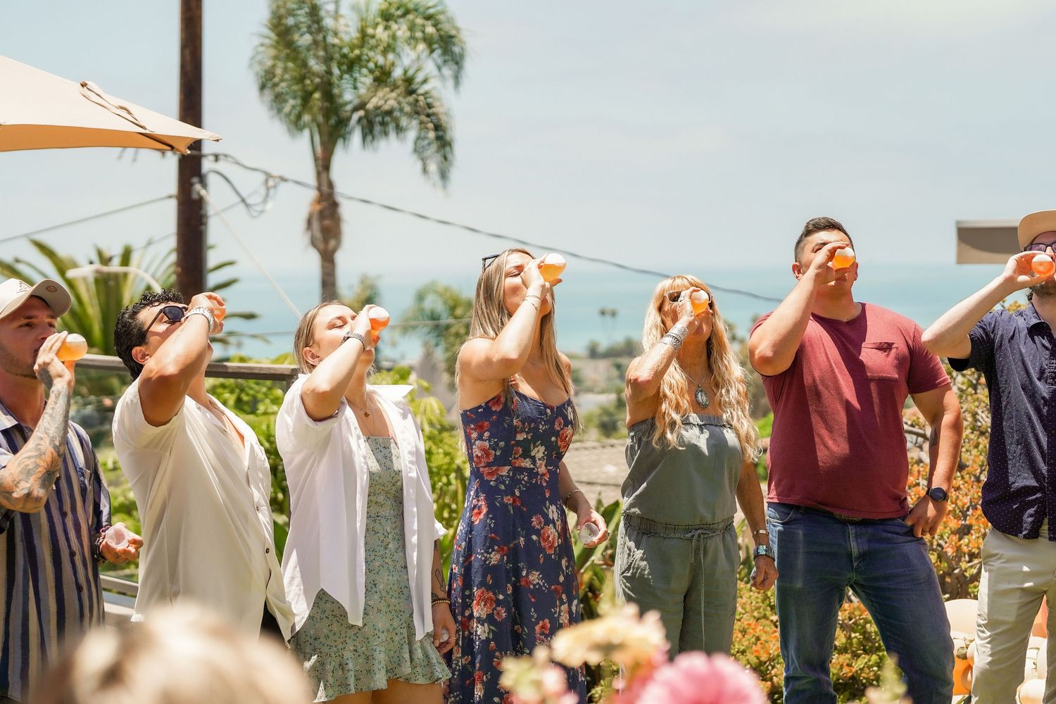 Group of friends raise small shot glasses in a toast at an outdoor terrace party with palm trees and ocean views in the background.