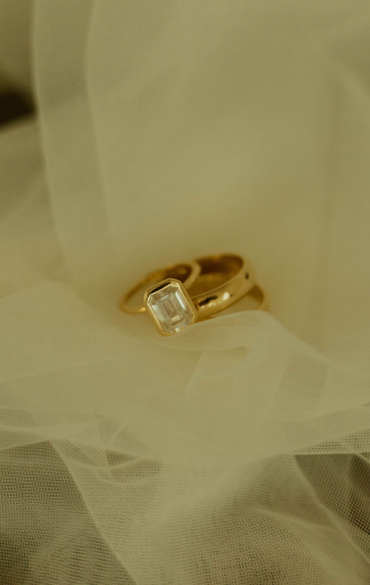 Gold emerald-cut diamond solitaire ring stacked with a gold band resting on soft ivory tulle fabric.