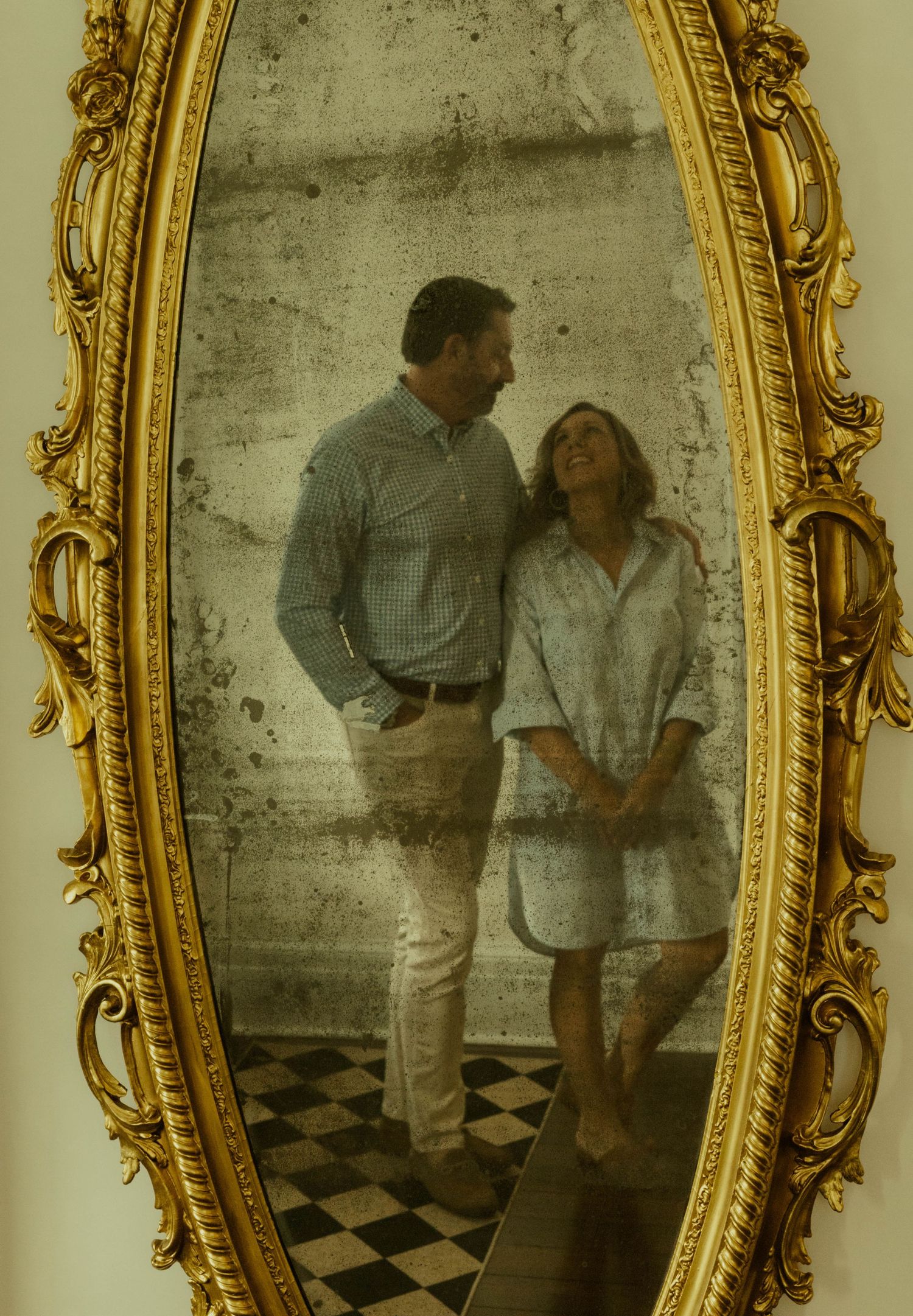 Smiling couple embracing is reflected in an ornate gold-framed antique mirror above a black and white checkered floor.