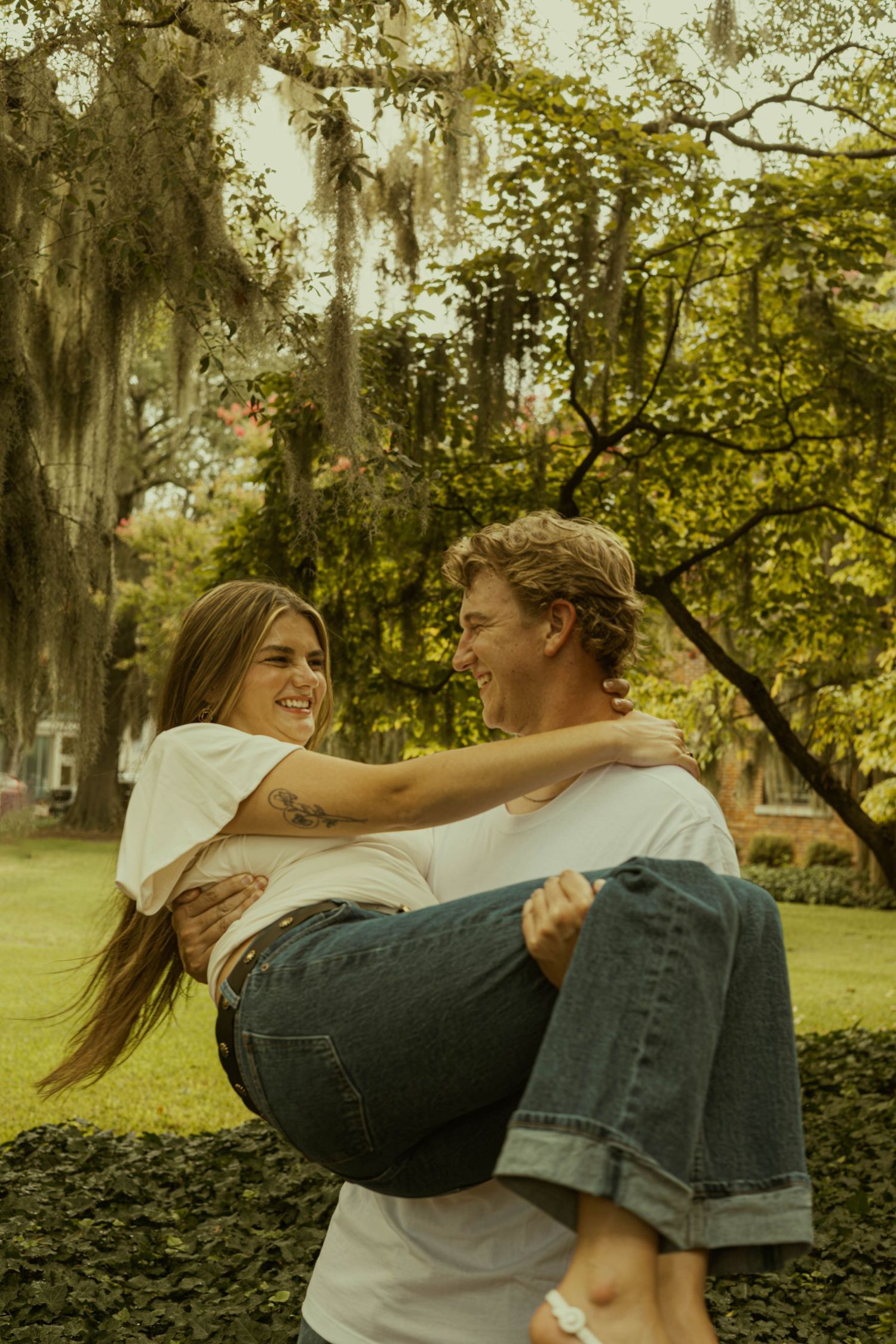 Smiling man carries his fiancée in his arms among moss-covered trees during a joyful engagement photo session.