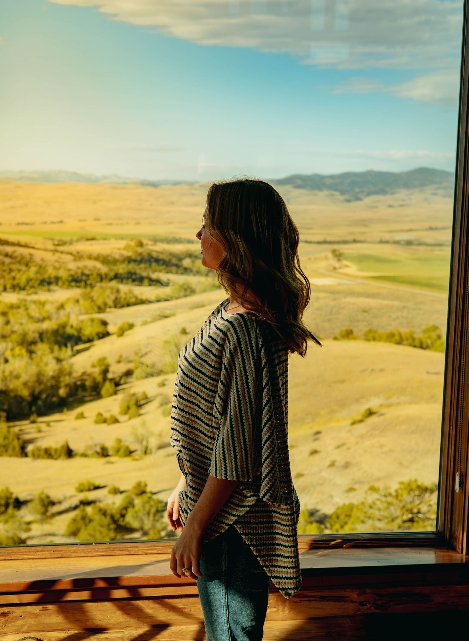 Woman in a striped top and jeans stands by a large window gazing at golden rolling hills and valley landscape under a blue sky.