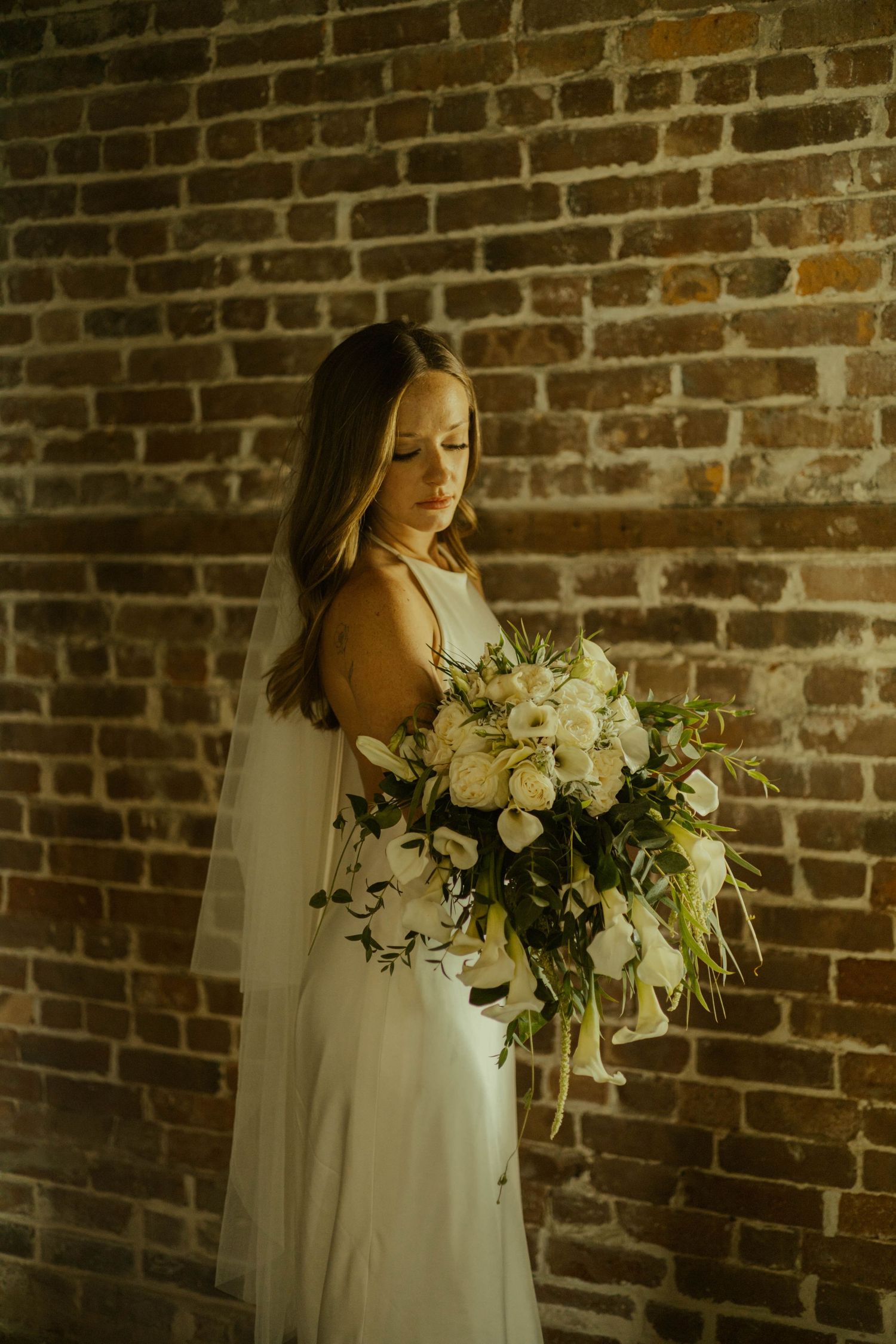 Bride in a white gown and veil holds a white floral bouquet while standing against a rustic brick wall.