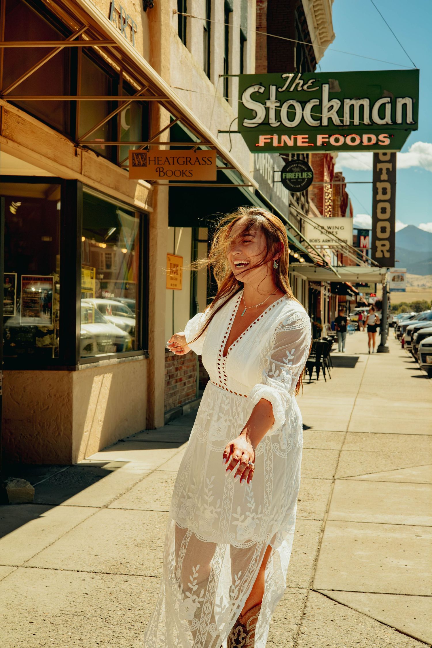 A woman in a flowing white lace dress laughs on a sunny sidewalk beneath The Stockman Fine Foods sign in a mountain town.