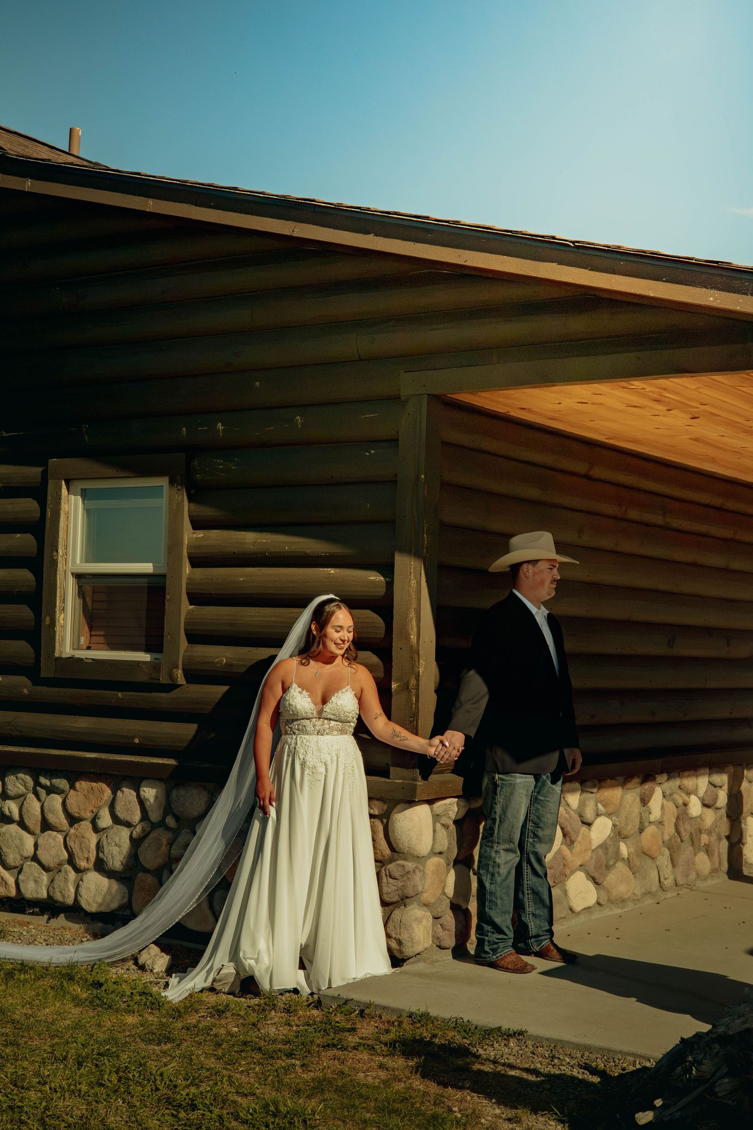 Bride in a flowing gown holds hands with a man in a cowboy hat outside a rustic log cabin with a stone foundation.