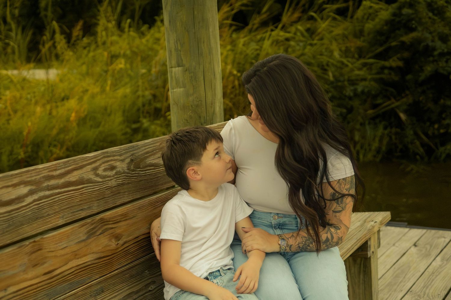 A dark-haired woman and a young boy in a white tee sit close together on a wooden dock bench by the water.