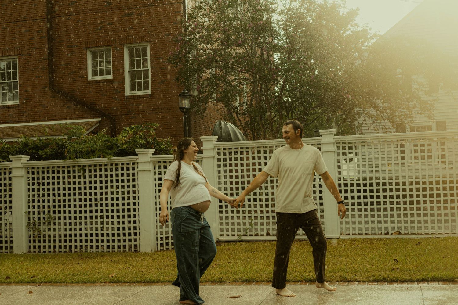 Pregnant woman and her partner hold hands while walking along a sidewalk in front of a white lattice fence and brick house in warm light.