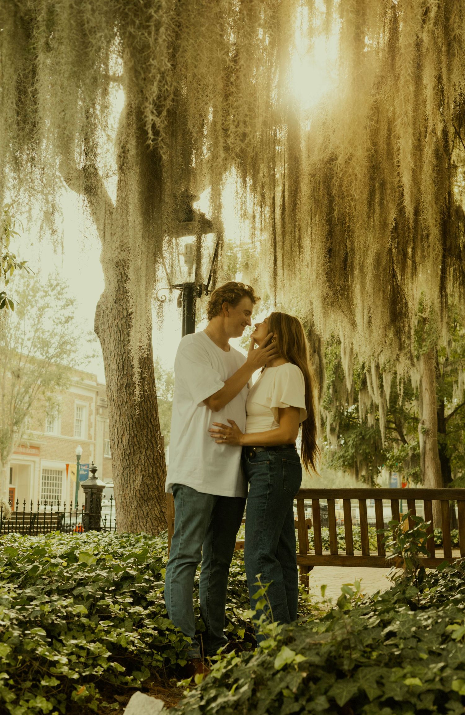 Engaged couple shares a tender moment beneath a moss-draped oak tree in a sunlit Savannah-style park.