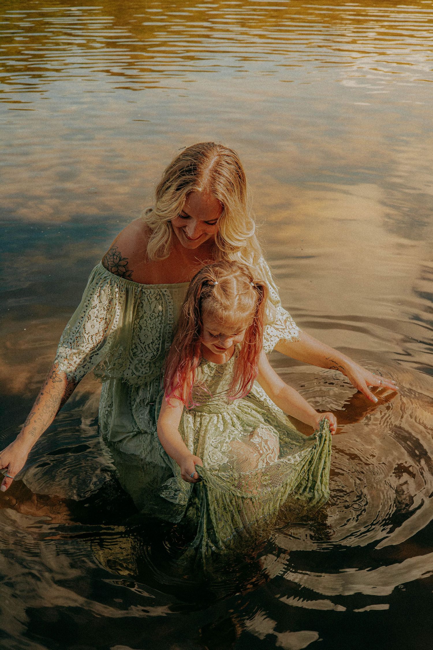 Smiling mother and young daughter with pink-tinted hair wade in golden-lit water wearing light green dresses.