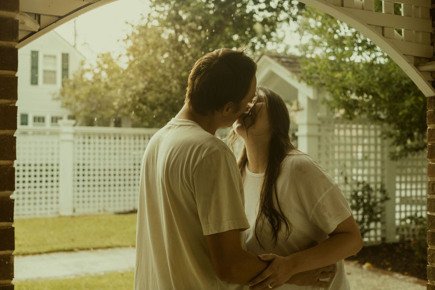 Couple kiss tenderly on a porch beside a white lattice fence in soft, warm light during their maternity session.