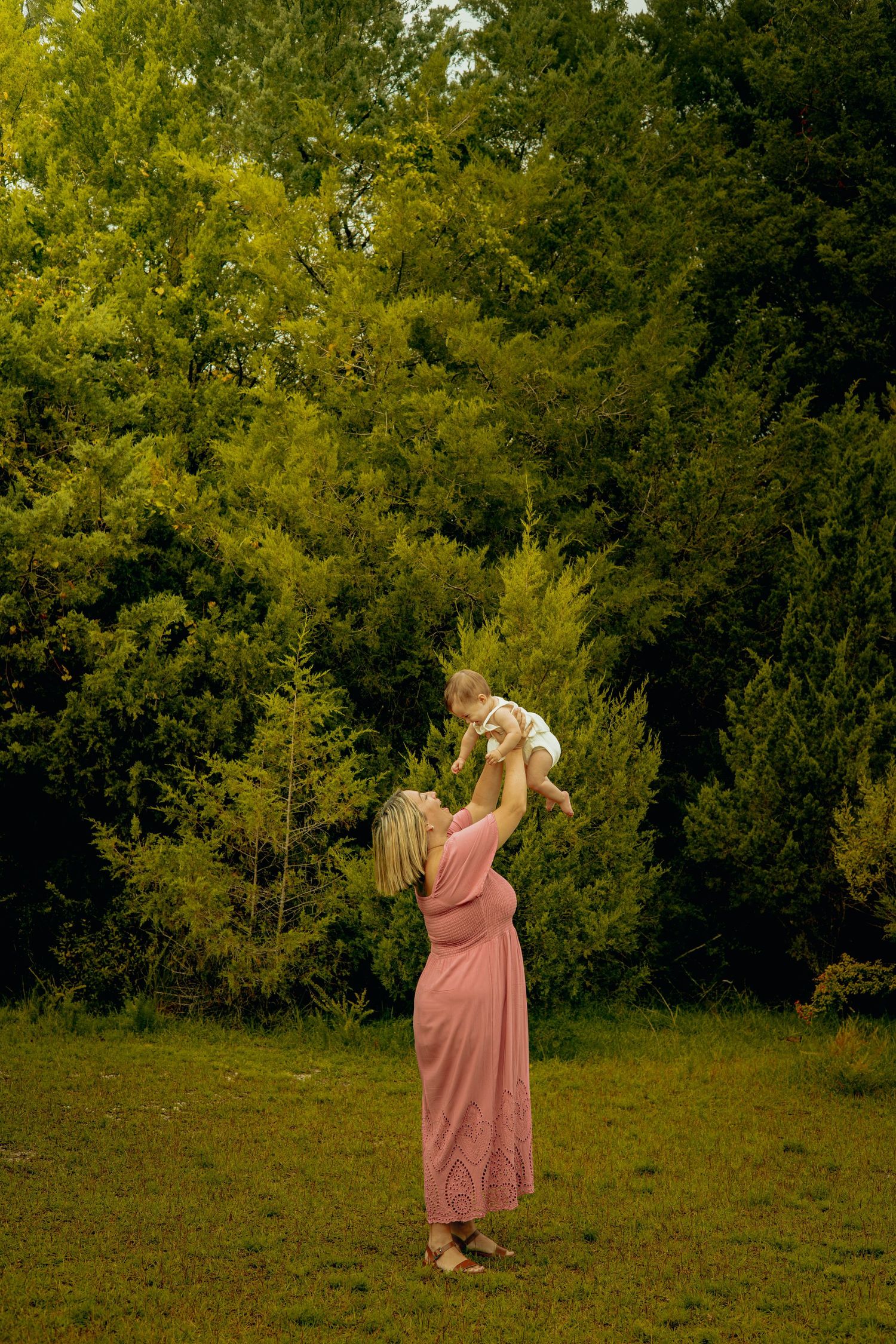 A mother in a long pink dress lifts her baby high into the air on a grassy lawn in front of lush green evergreen trees.