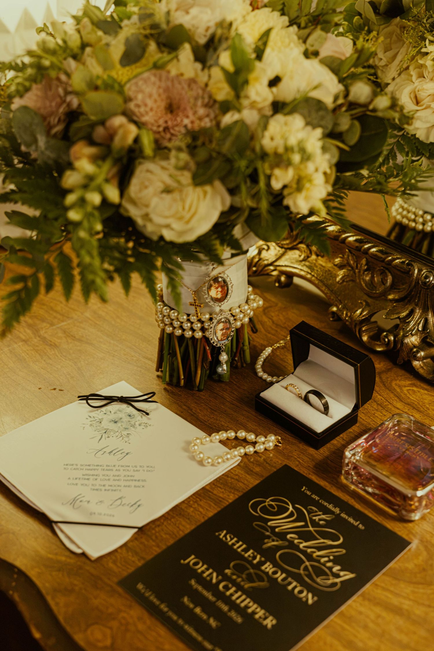 Wedding details on a wooden table including a bouquet, black invitation, pearl jewelry, perfume, and a ring in a box.