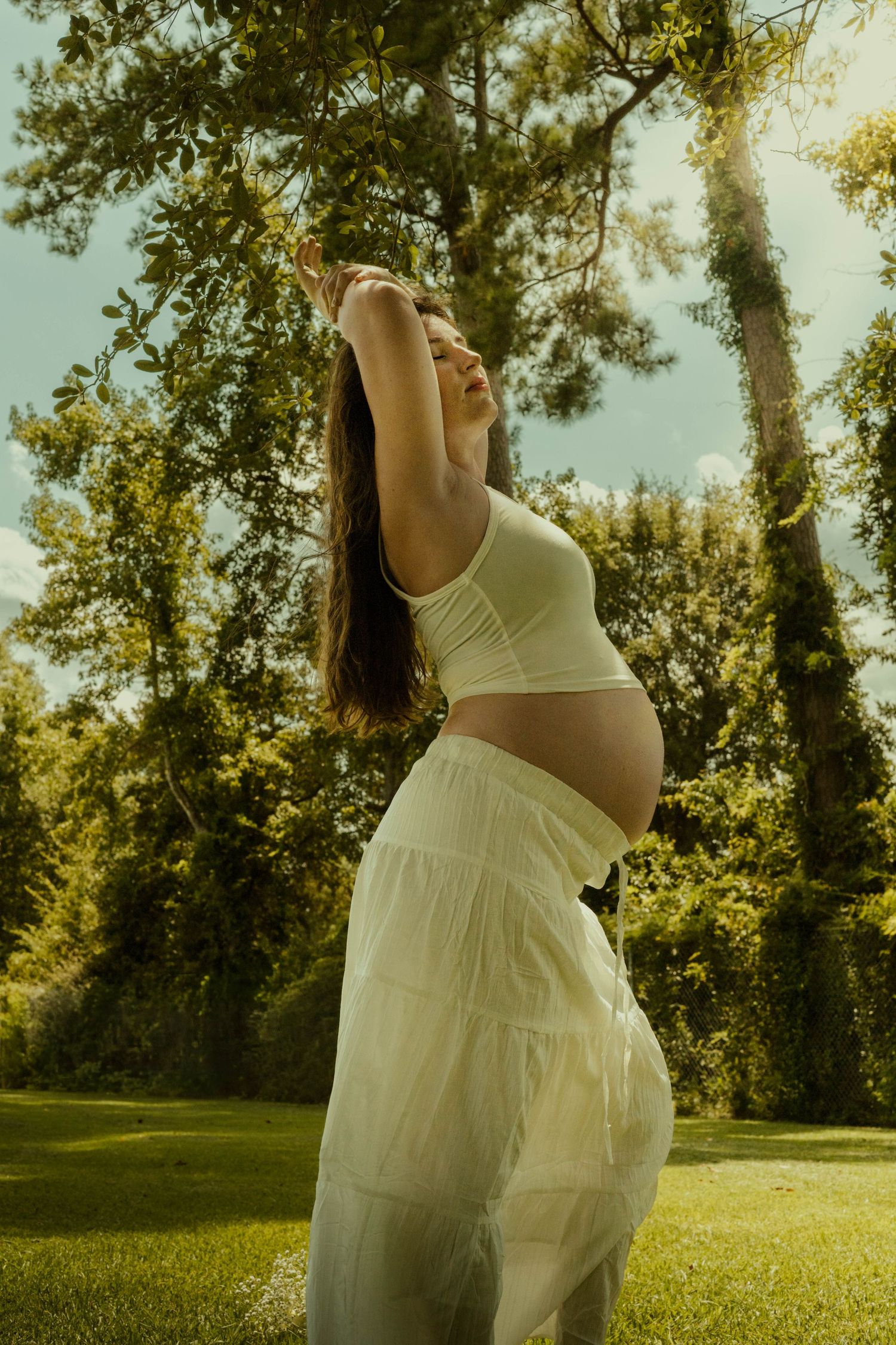 Pregnant woman in a white skirt and crop top stretches her arms overhead on a sunlit lawn surrounded by lush green trees.