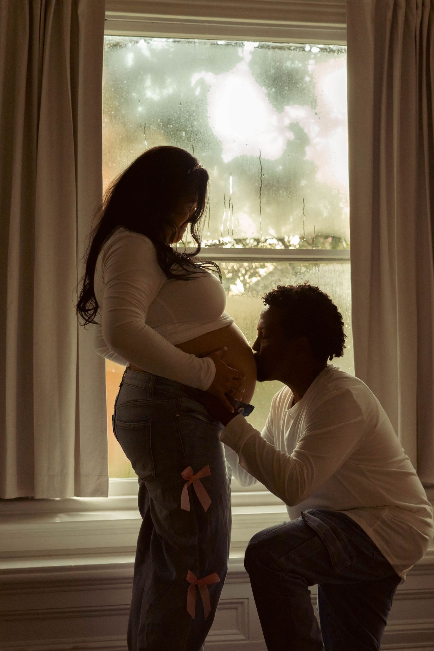 Silhouetted man kneels to kiss his pregnant partner's belly beside a rain-streaked window in a moody maternity portrait.