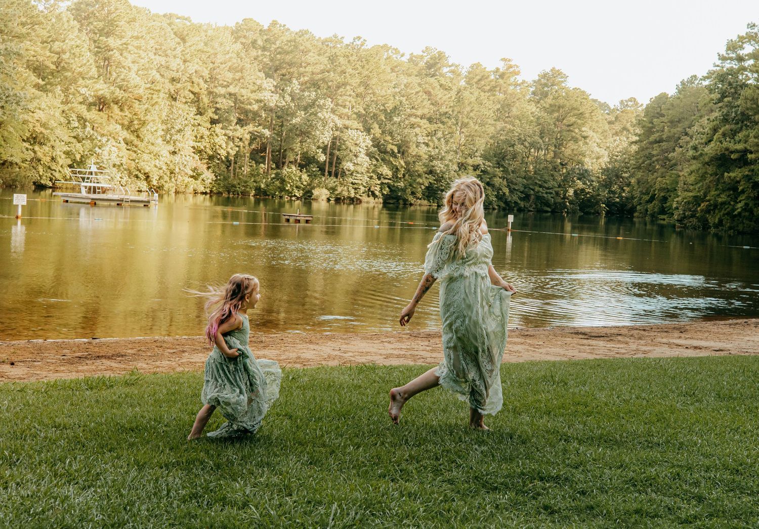 Woman and young girl in matching green dresses run across a grassy lawn beside a calm lake surrounded by tall trees.