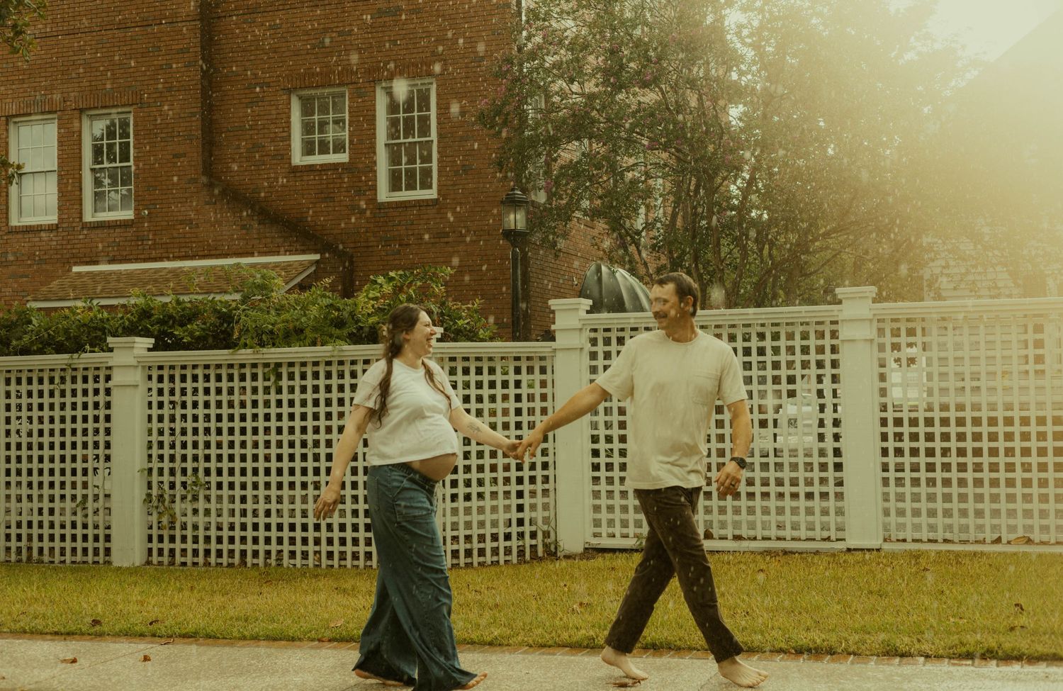 Expecting couple holds hands and walks along a sidewalk past a white picket fence and red brick house in warm sunlight.