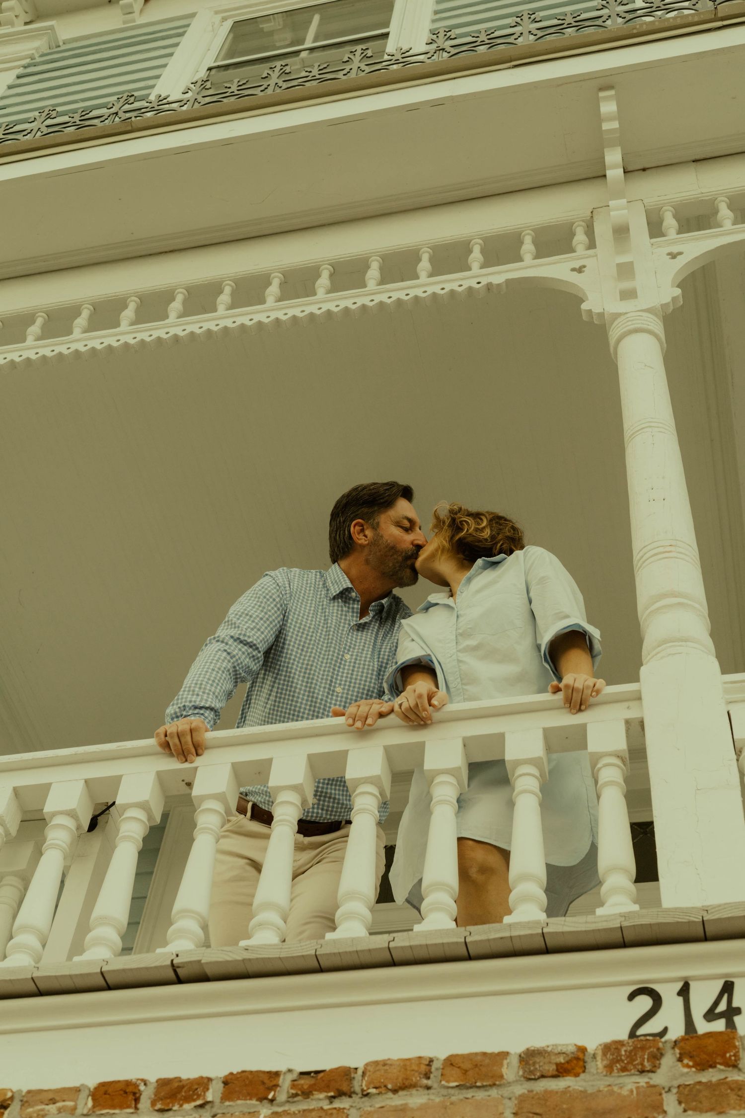 Couple kiss on a white-railed balcony of a historic Southern home, photographed from below during an engagement session.