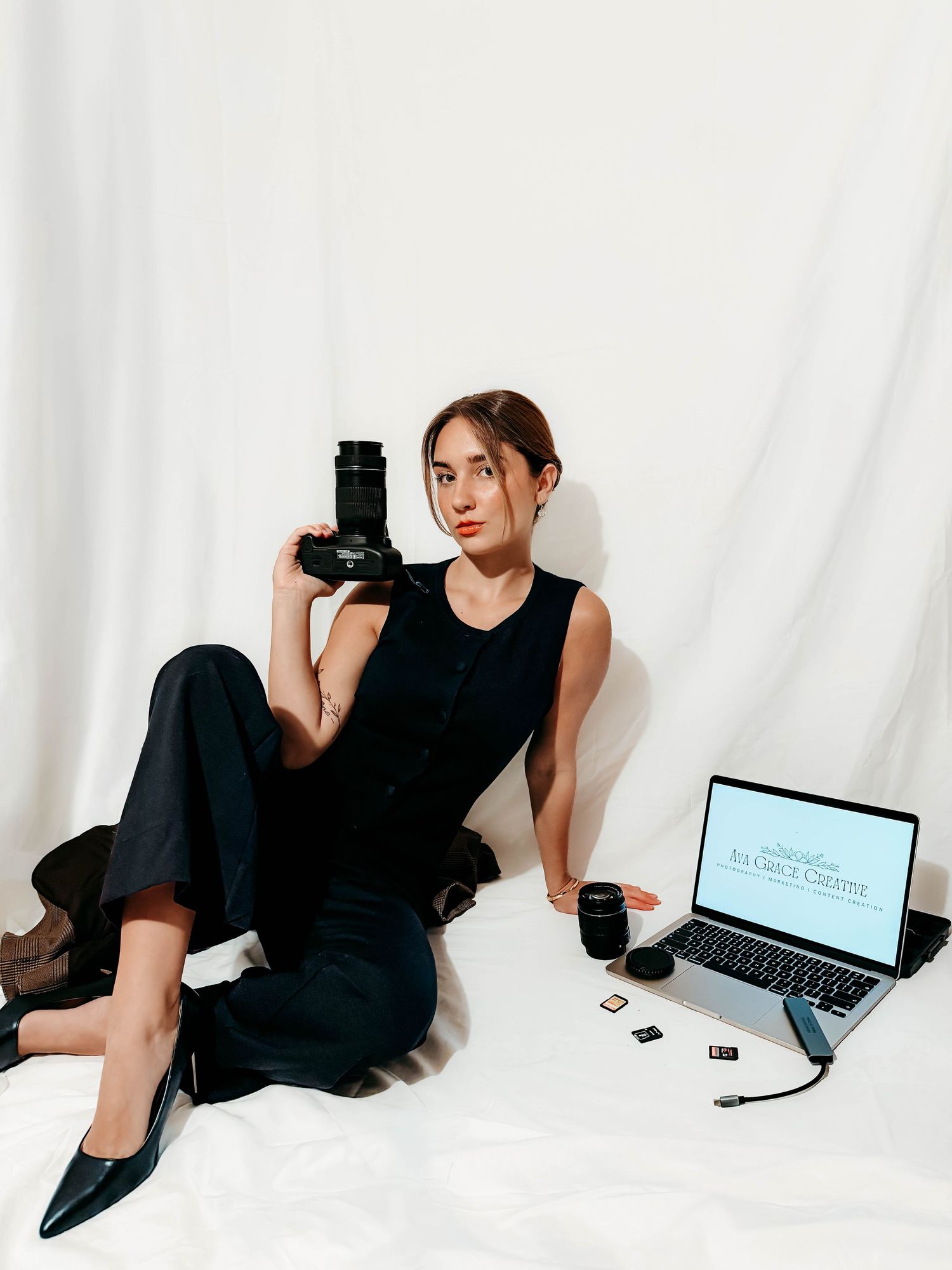 Photographer in black outfit sits on a white backdrop holding a camera beside an open laptop, lens, and memory cards.
