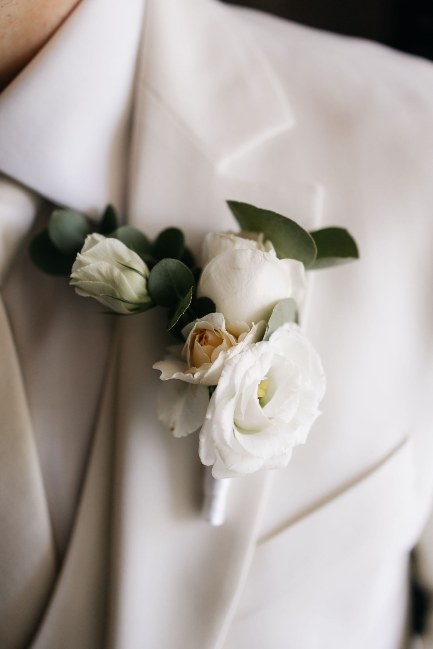 Close-up of a white rose and eucalyptus boutonniere pinned to the lapel of a cream wedding suit jacket.