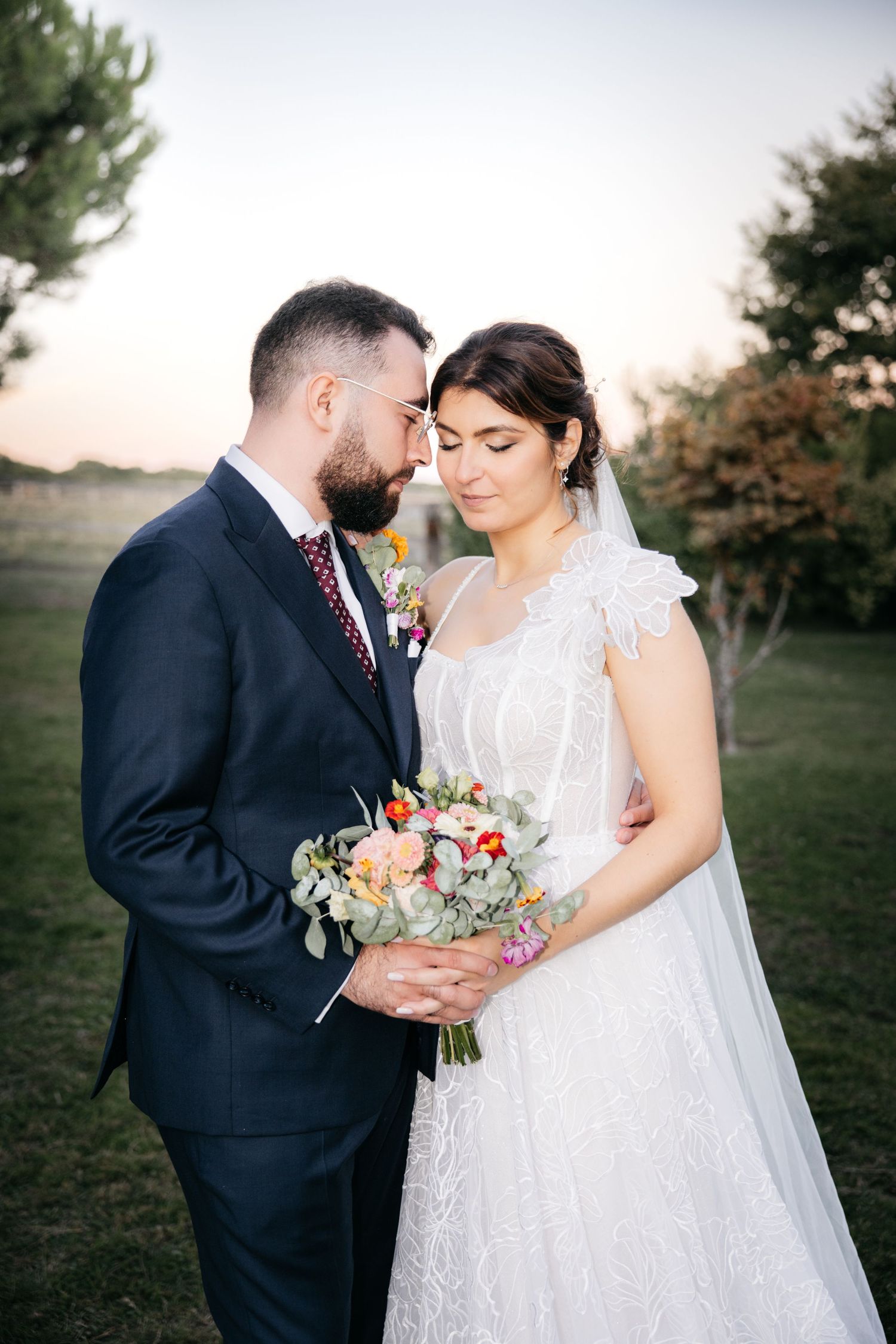 Bearded groom in a navy suit embraces a bride in a lace gown and veil holding a colorful bouquet in a field at sunset.